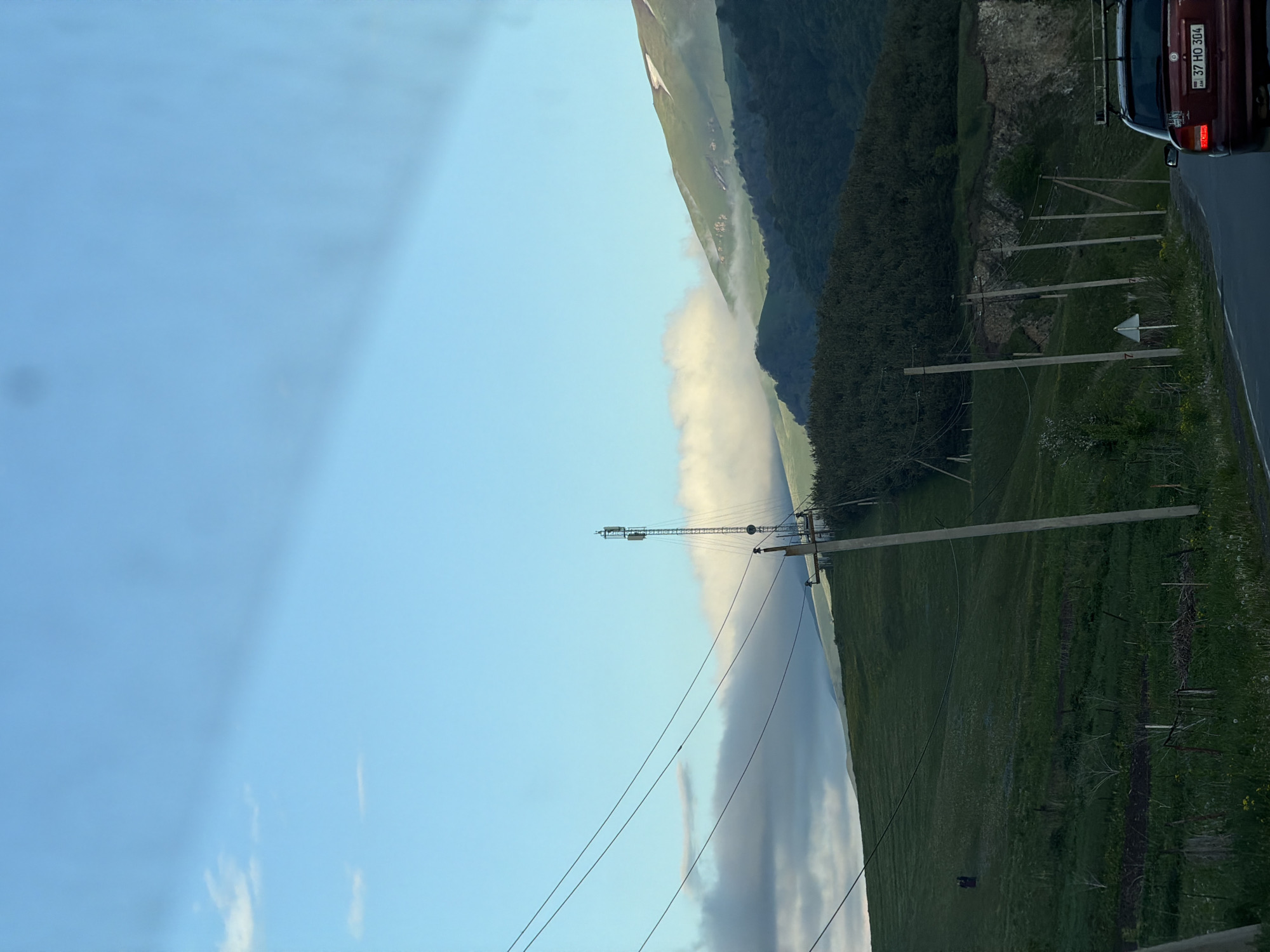 The image depicts a mountainous landscape in Fioletovo, Armenia, partially obscured by a low-lying cloud formation. The sky is blue with some light clouds. The land is green with vegetation, and power lines run along the roadside. A red vehicle is visible on the road in the upper right corner, with license plate number "37 HO 304." Tall, wooden utility poles stand along the right side of the image.