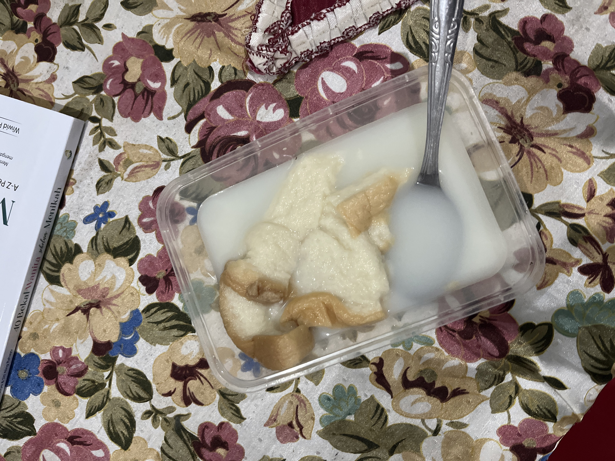 A close-up, overhead shot shows a clear plastic container filled with a white, milky liquid and pieces of bread. A silver spoon rests inside the container, partially submerged in the liquid. The container is placed on a floral-patterned tablecloth featuring large pink, yellow, and blue flowers with green leaves. In the upper right corner, a white book with a title partially visible is propped up against something off-camera. The book's title includes "40 Bekal Wanita sebelum Menikah." The scene is indoors, likely on a table. There is no discernible connection to the city of Glagah Kulon, Indonesia.