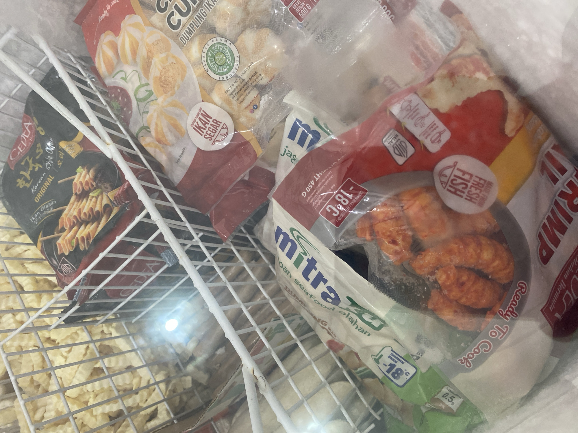 The image shows a chest freezer filled with various frozen food items. In the foreground, a clear plastic bag contains several pieces of orange, breaded food, labeled "Ready To Cook FISH FRESH." Behind this, partially obscured, is a bag of what appears to be shrimp or fish dumplings, with text including "GULAI." To the left, a white bag with green and blue lettering reads "mitra oah seafood olahan." In the upper right, a black package features Korean text and an image of rolled food items, with the brand "CEDA" visible. Next to it, a red and white bag shows images of round, striped food items. In the background, a white wire basket holds a large quantity of crinkle-cut fries. The overall environment appears to be a commercial freezer unit, likely in a market or food establishment in Payatieng, Indonesia, given the visible Indonesian text on several packaging labels. The lighting suggests an indoor setting with some glare from the freezer's interior light. There are no people or animals visible in the image.