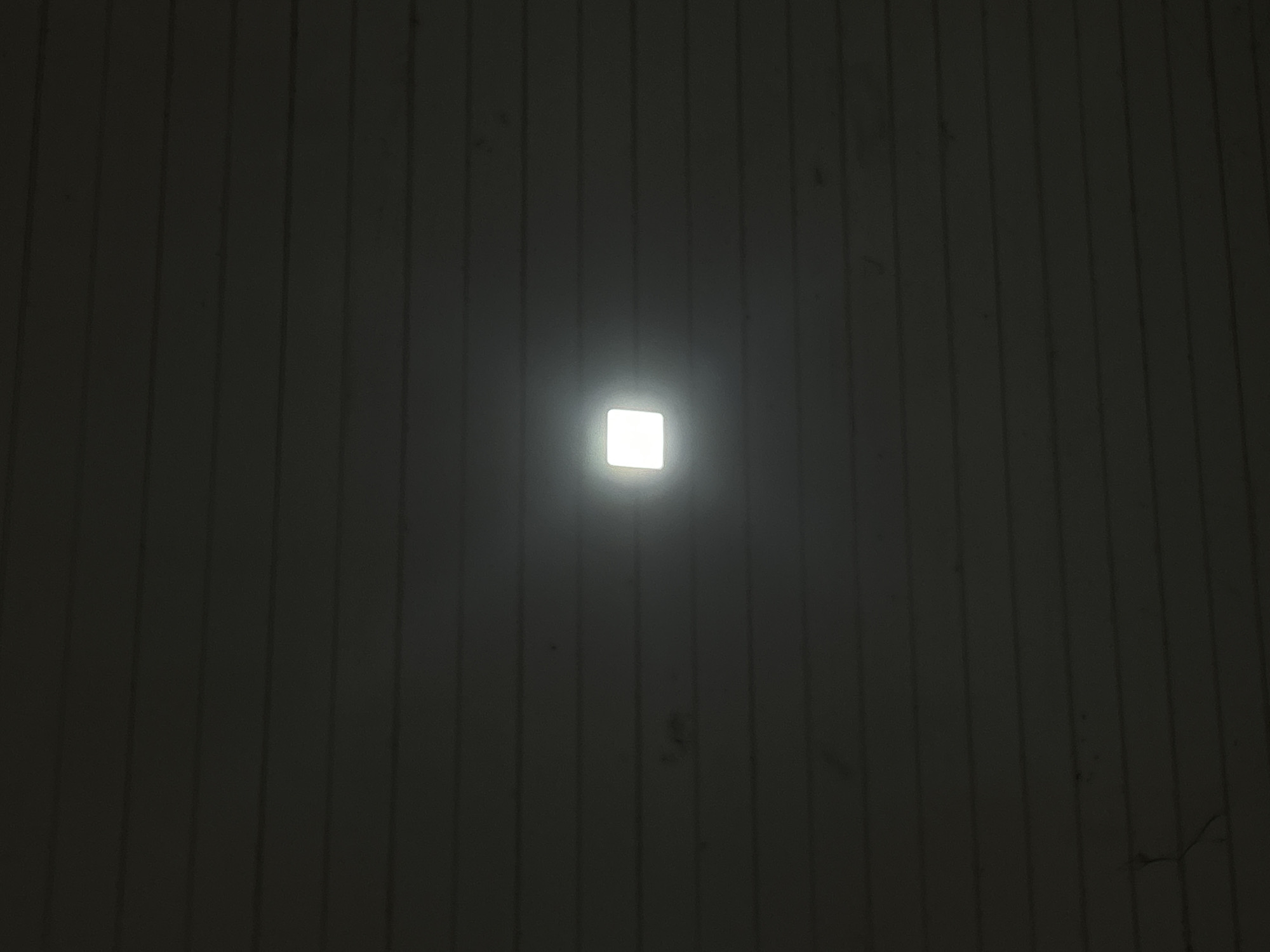 The image presents a dark, interior scene dominated by a single, bright, square-shaped light fixture centrally positioned. This fixture emits a strong white light, creating a distinct glow against the otherwise dim surroundings.
The background appears to be a ceiling or wall, characterized by a pattern of dark vertical lines, suggesting paneling or slats, possibly made of wood or a similar material. Some faint smudges or dirt marks are visible on these dark surfaces, along with minor imperfections such as a possible scratch or crack. The overall darkness of the environment suggests the photograph was taken at night or in a room with minimal or no natural light, with the central fixture serving as the primary light source.
There are no people or any specific activities depicted in the image. No text is visible. The scene is devoid of strong emotional cues, presenting a straightforward view of an illuminated architectural detail. The location context for this scene is Reuleut Timu, Indonesia.