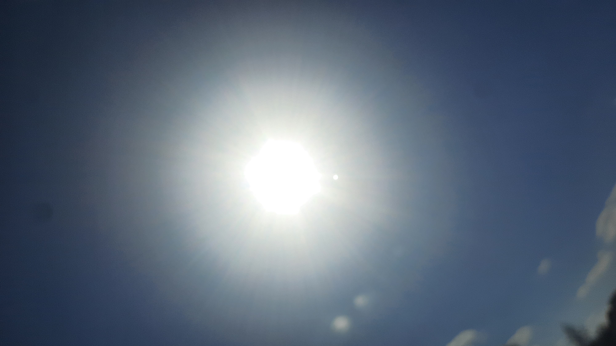 The image shows a bright sun in a clear blue sky, with some lens flares present. The sun is positioned in the upper central part of the frame. There are no people, objects, or specific landmarks visible that would indicate a location within the city of Шклоў, Belarus. The scene is a generic depiction of a sunny sky.