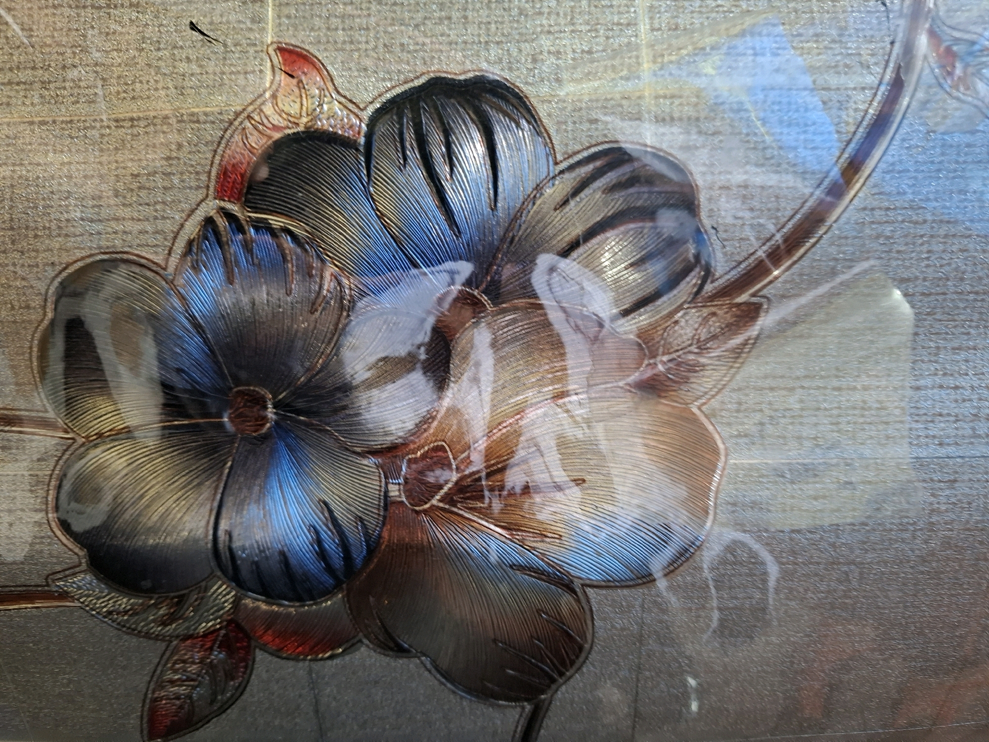 The image depicts a close-up of a decorative glass panel featuring embossed floral motifs. The design consists of stylized flowers with prominent petals, rendered in metallic tones of blue, brown, and red, with fine linear textures on the petals. The background has a textured, possibly tiled or wallpapered, appearance. Light reflects off the glass, creating bright highlights and obscuring some details. There are no people or any recognizable landmarks of Sebong Lagoi, Indonesia visible in the image. The scene is entirely focused on the decorative glass artwork.