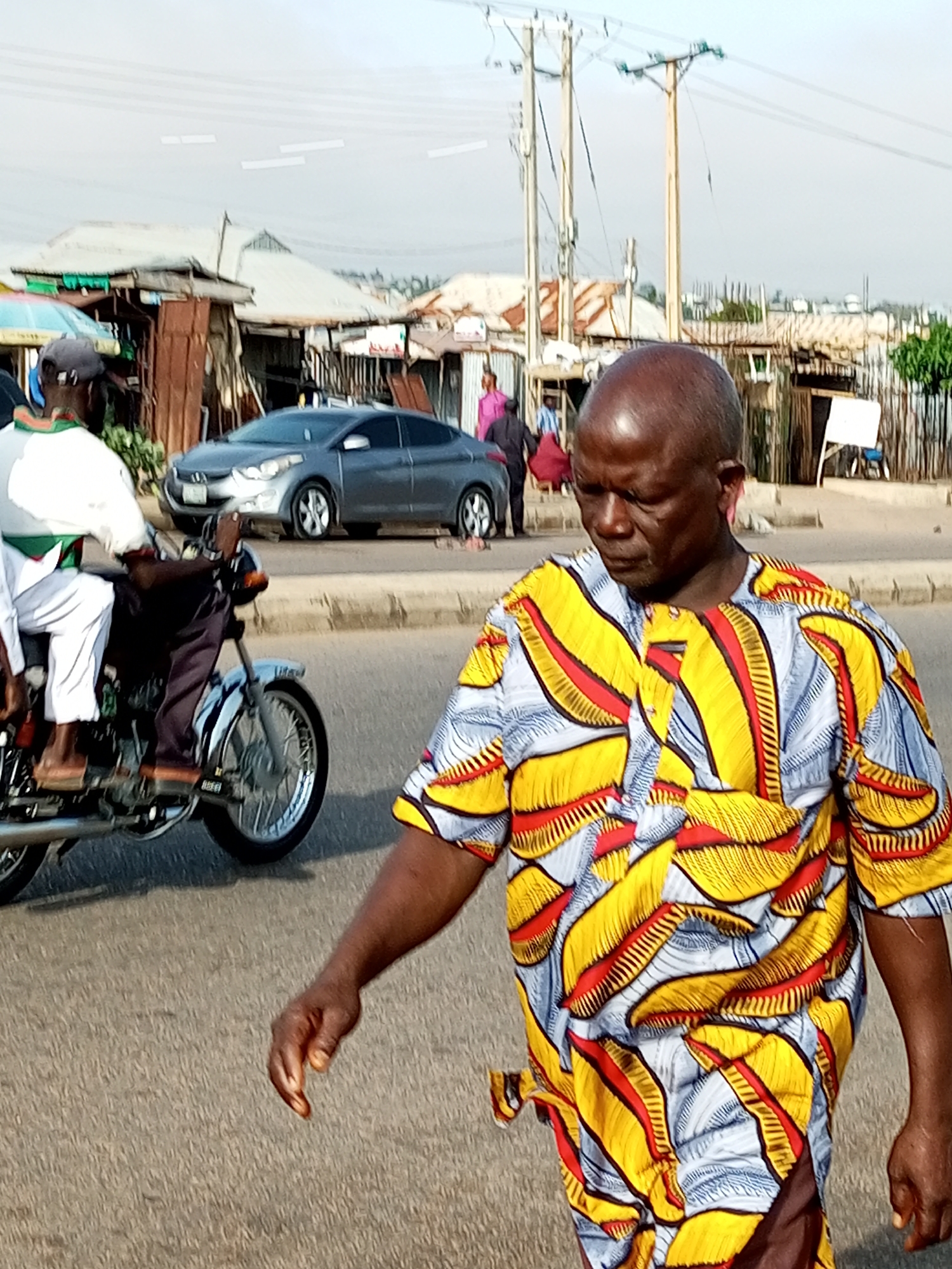A medium shot shows a bald, dark-skinned man walking on a street in Tempure, Nigeria, wearing a brightly colored, patterned shirt with yellow, red, and blue designs. He is in the foreground and looking down. Behind him, a person on a motorcycle wearing a white and green garment is visible. In the background, there is a silver car, some corrugated metal structures that appear to be market stalls, and utility poles with wires. The street is paved, and there is a curb. The sky is overcast.