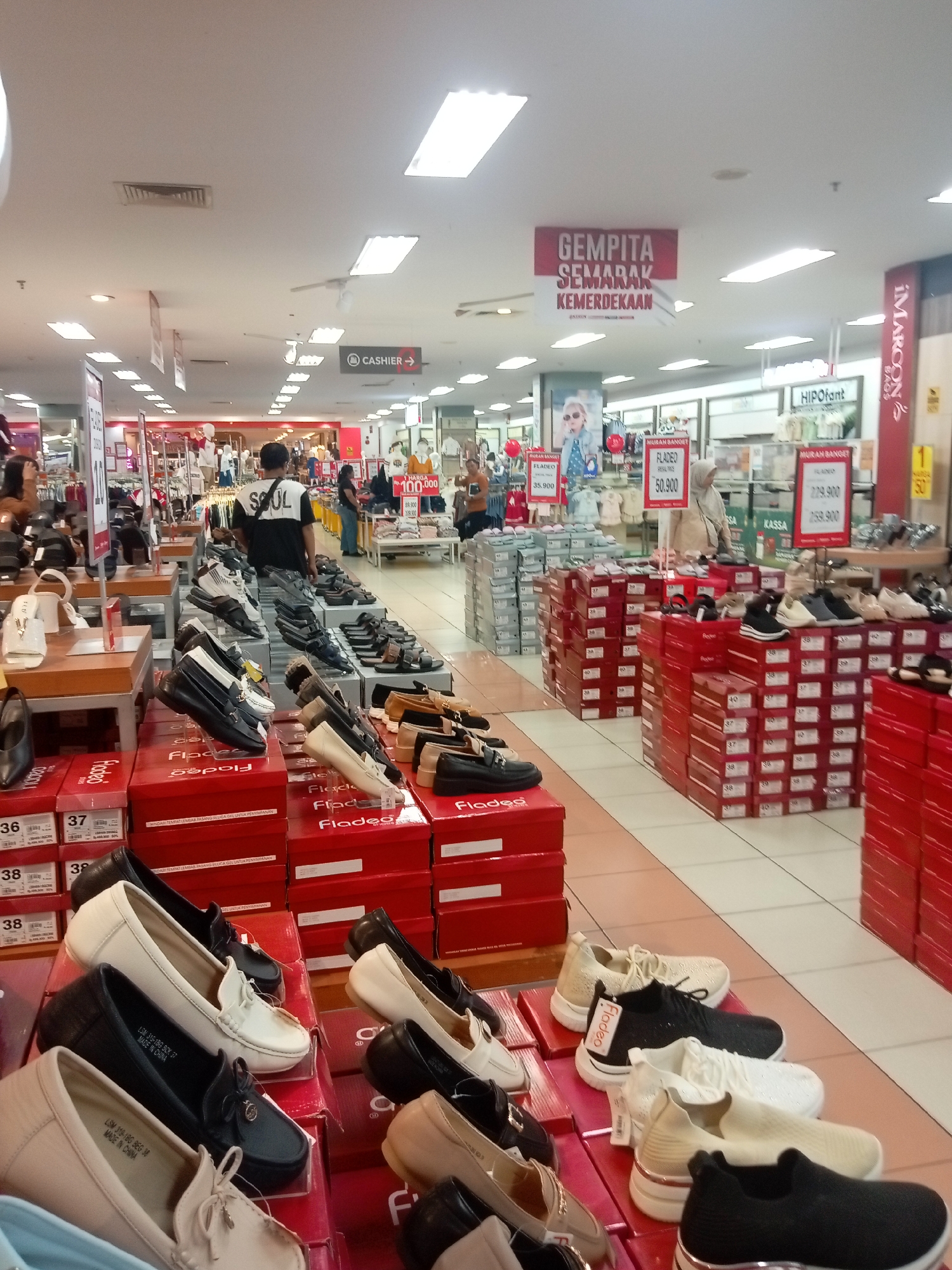 The image depicts the interior of a shoe store, likely located in Batam, Indonesia, given the Indonesian language visible on signs. The store is well-lit with overhead fluorescent lighting and features rows of shoe displays. Various types of footwear, including women's heels, flats, sneakers, and men's sandals, are arranged on shelves and stacked boxes. Several price tags and promotional banners are visible, with text such as "MURAH BANGED" (very cheap) and "GEMPITA SEMARAK KEMERDEKAAN" (celebrate the spirit of independence) indicating sales or events.
In the foreground, a display of women's flats and loafers in various neutral colors like beige, white, and black are prominently featured. Behind them, more shoe boxes are stacked in neat rows, predominantly in bright red. Further back, the store opens up to reveal more shoe displays, clothing racks with mannequins, and various sections of the store.
Several individuals are present in the scene, including shoppers browsing the merchandise and store staff attending to customers or arranging displays. One person in the middle ground is wearing a t-shirt with "SOUL" printed on it. Another person is seen walking towards the back of the store, and others are scattered throughout the aisles. A cashier counter is also visible in the background. The overall atmosphere suggests a busy retail environment.
