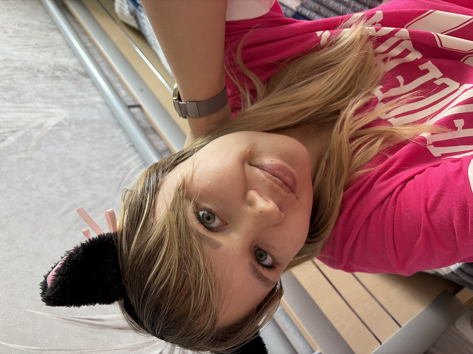 A woman with blond hair and green eyes is facing the camera while wearing cat ears. She is wearing a pink shirt and a watch. She appears to be lying down, and a bed frame is visible behind her. The location is Chaikovsky, Russia.