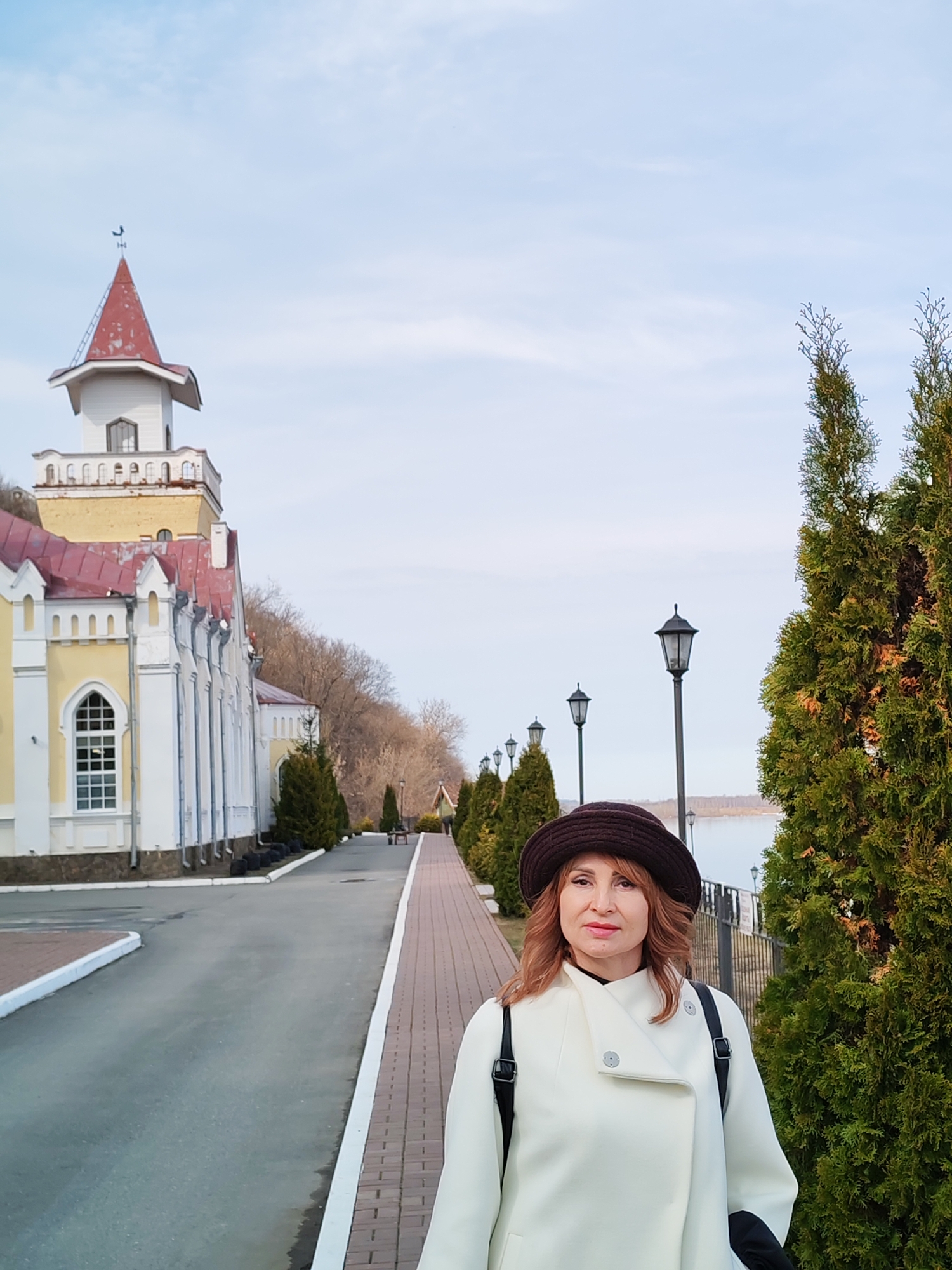 The image captures a woman standing on a brick pathway in Sarapul, Russia. She is facing the camera, wearing a white coat and a brown hat. She has a backpack on. To her right are tall, evergreen trees.
To the left is a yellow and white building with a red roof and a distinctive tower. A road runs adjacent to the building, lined with trees and lampposts. In the background is a body of water. The sky is overcast.