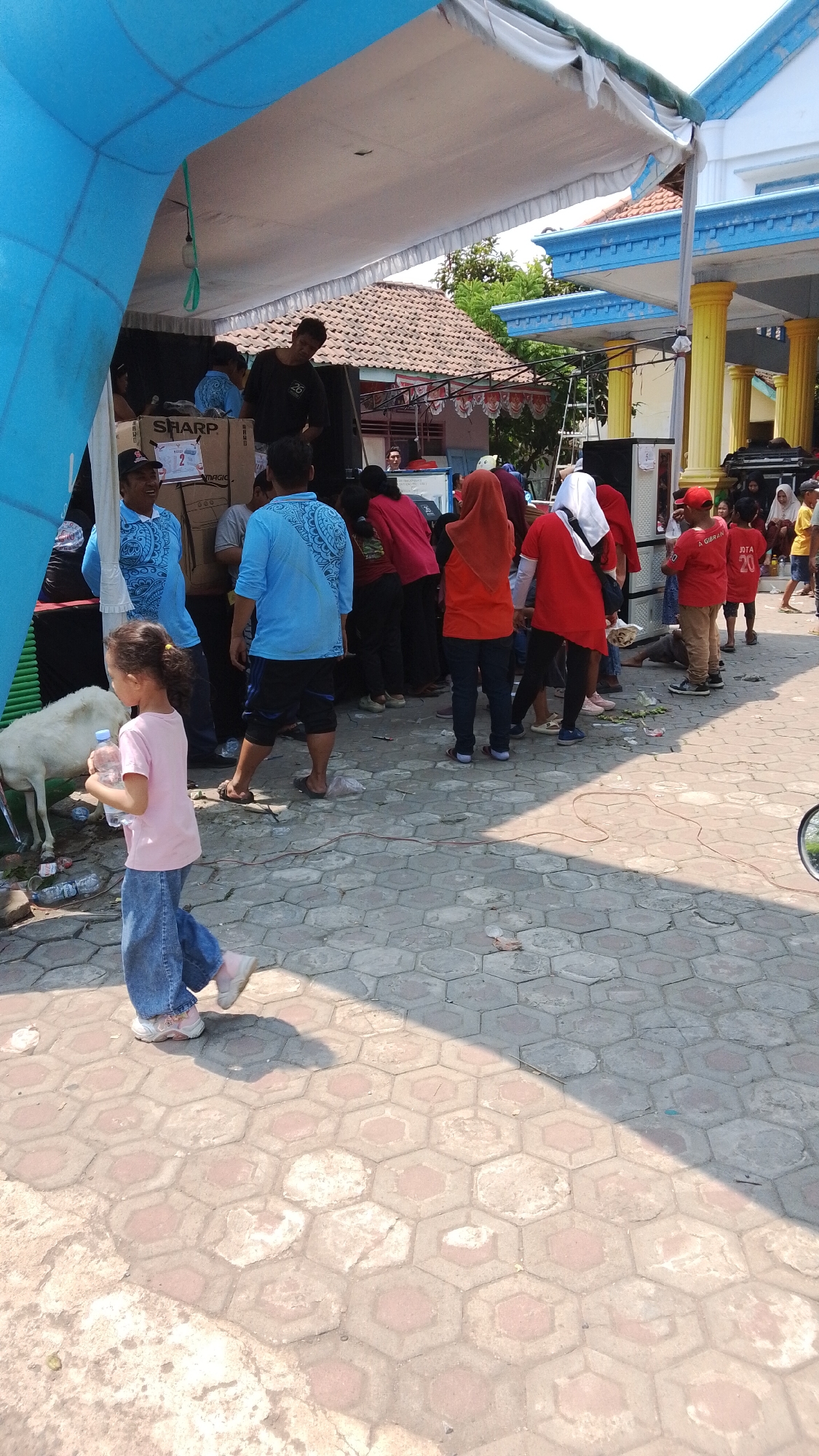 The image depicts an outdoor scene in what appears to be a public gathering or market setting in Mancar, Indonesia. The foreground is dominated by a paved hexagonal stone walkway, with patches of sunlight and shadow indicating a bright, sunny day. A young girl in a pink t-shirt and denim jeans walks from left to right, holding a plastic bottle of water. To her left, a white goat stands near some discarded items.

Behind the girl, a group of people are gathered under a large blue and white tent structure. Some individuals are wearing blue shirts with a patterned design, while others are in red shirts. There are boxes and a sign that says "SHARP" visible within the tent. The background shows buildings with traditional Indonesian architecture, including tiled roofs and blue accents on the building on the right. Several trees and greenery are also visible, suggesting a suburban or semi-rural environment. The overall impression is a community event or a bustling marketplace.