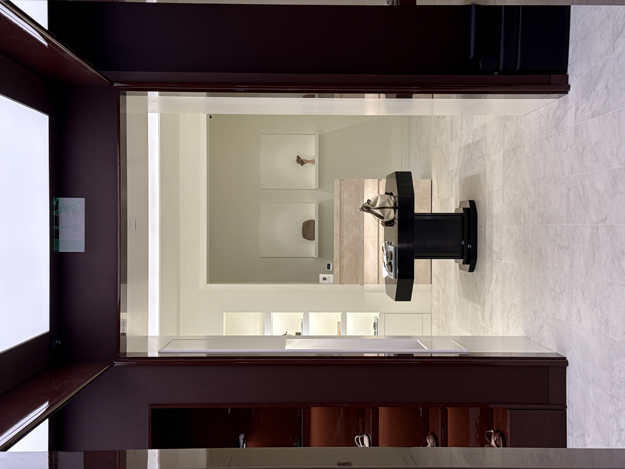 The image captures an interior view, seemingly a store or showroom in Perm, Russia. The perspective is looking through a doorway or archway, framed by dark reddish-brown trim. The interior features a light, neutral color palette with what appears to be marble or stone flooring. In the center of the view is a black pedestal table with a bag or purse on top of it. The background includes displays and shelves, with minimalist decor such as a framed image of a hand. The overall impression is one of modern, upscale design.