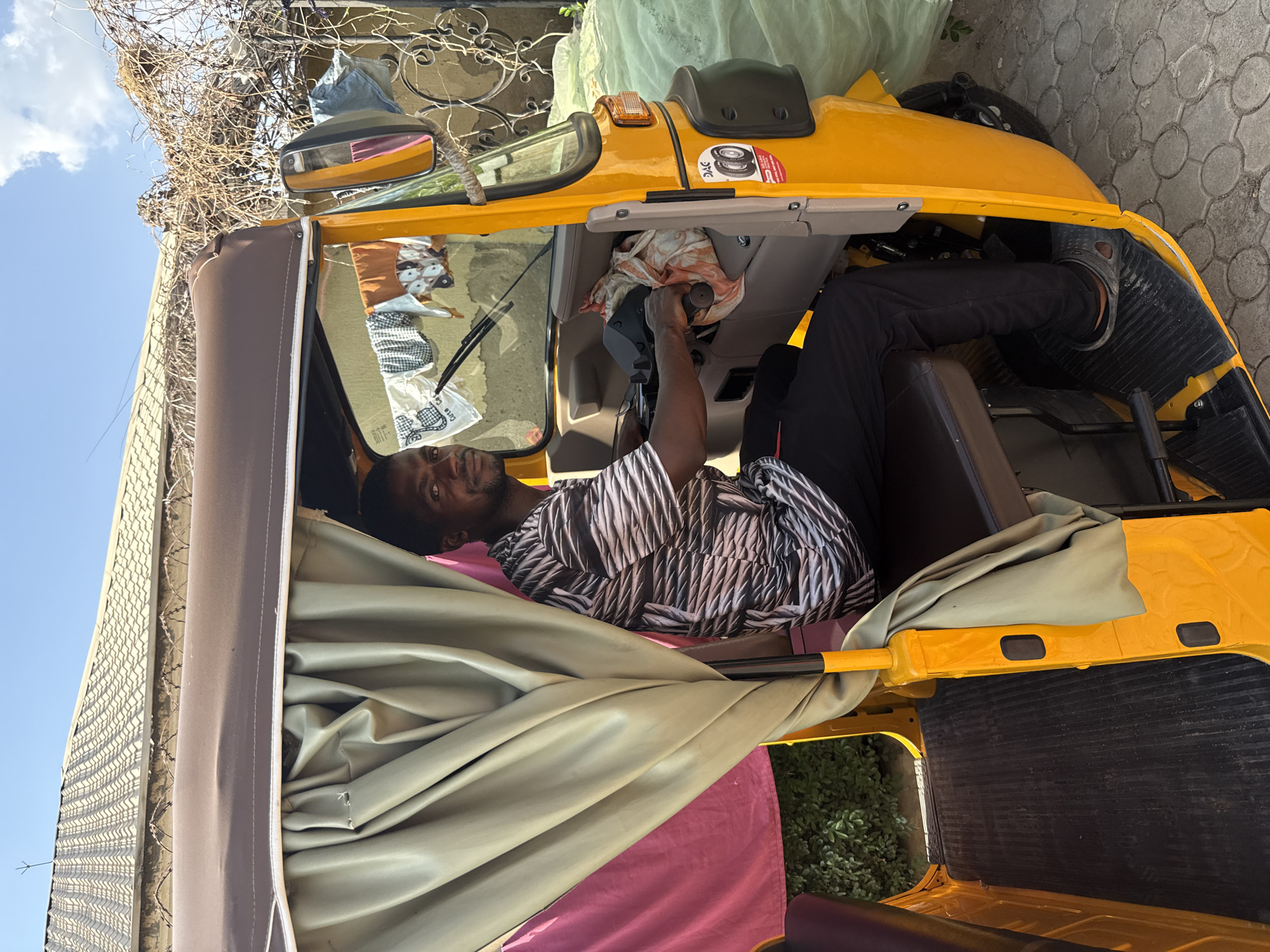 The image captures a relaxed moment featuring a man resting inside a yellow three-wheeled auto-rickshaw, commonly known as a "Keke Napep" in regions like Abuja, Nigeria. The scene is set outdoors during bright daylight under a clear blue sky with sparse white clouds.
The primary subject is a man, appearing to be in his twenties or thirties, with a light beard or stubble. He is dressed in a patterned shirt with a black and white or grey geometric design. He is lying down on his side inside the vehicle, with his head propped up and looking directly towards the camera with a neutral and calm expression. One hand is visible, gripping a component near the steering column, which is covered with a light orange and white patterned fabric or rag. His legs, clad in dark trousers and open-toed shoes (possibly Crocs), are visible extending towards the front of the vehicle.
The vehicle itself is distinctly yellow, with a soft, retractable roof that appears to be brown or tan. Details like a rearview mirror on the driver's side, a windshield, and parts of the vehicle's interior dashboard are visible. A small sticker with the letters "DAC" and an image of tires is affixed to an interior panel. In the background, inside the vehicle, a white plastic bag with the word "Care" partially visible can be seen.
The setting outside the vehicle includes a paved ground made of interlocking blocks, suggesting a street, parking area, or driveway. Beyond the vehicle, there's a light-colored building or wall topped with a fence featuring thorny or barbed wire, and some green foliage is visible near the bottom right of the frame. The overall atmosphere conveys a sense of a break or leisurely moment for the driver of the auto-rickshaw.