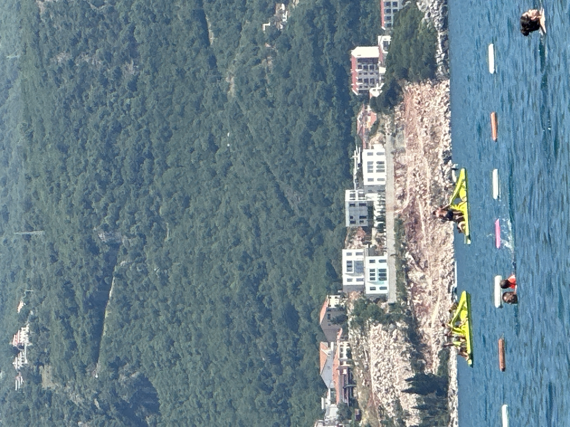 This is a vertical, medium-distance, eye-level shot of the coastline in Boreti, Montenegro. The foreground consists of calm, blue water with several floating buoys and a few people visible. In the middle ground, there are two yellow pedal boats with people on them. One pedal boat is closer to the shore and has several individuals, while another is further out with one person. Behind the boats, a rocky embankment is visible with modern white and red-roofed buildings situated on top of it. The background is dominated by a lush, green, forested hillside dotted with more houses. The sky is clear and blue, indicating a sunny day.