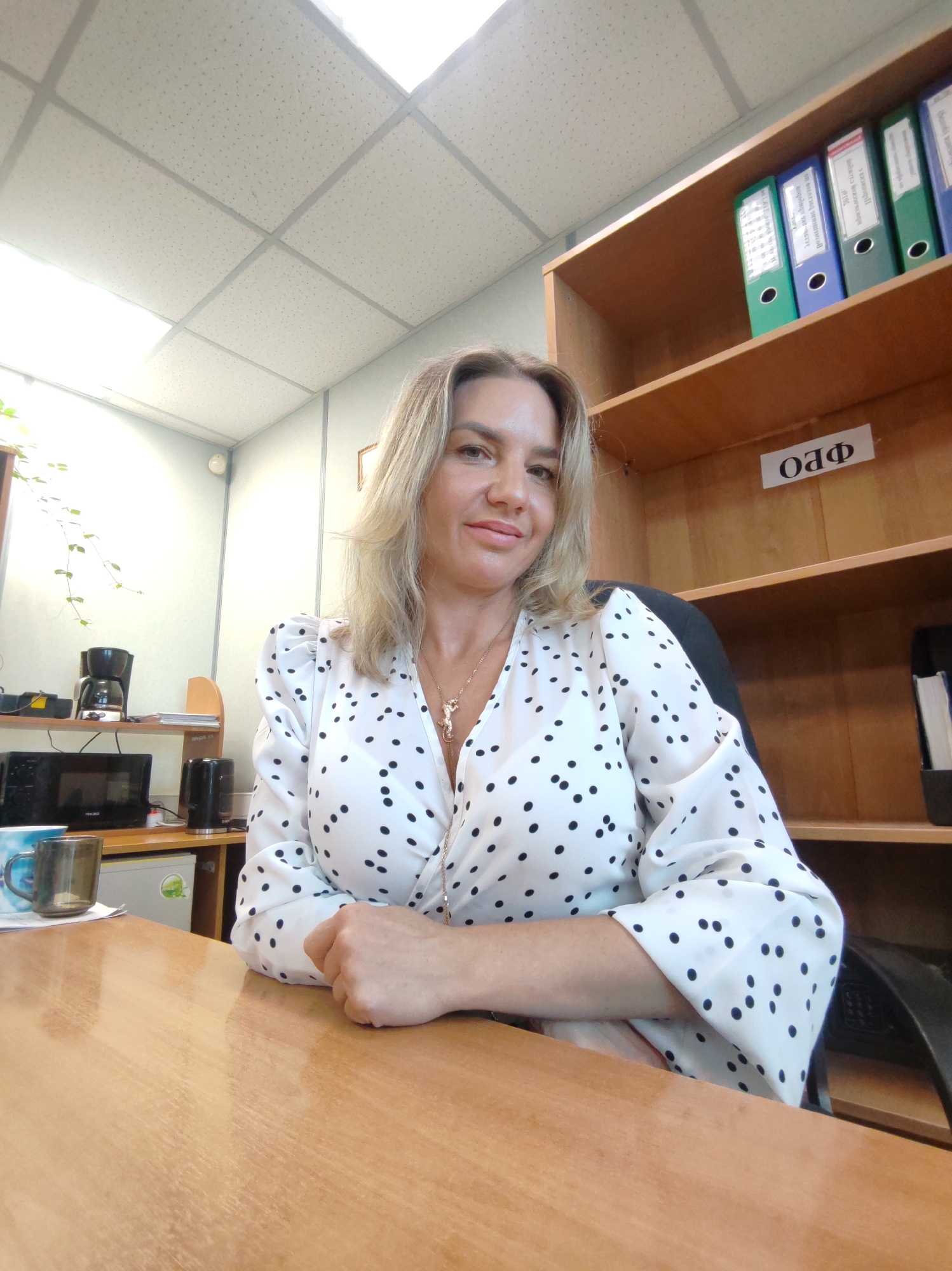 A woman is pictured from the chest up, seated at a light brown wooden desk in what appears to be an office or personal workspace. She has light-colored hair, a pleasant, soft smile, and is looking directly at the camera. She wears a white blouse adorned with black polka dots, featuring a V-neckline and somewhat puffy long sleeves. A gold-toned necklace with a detailed pendant, possibly an animal figure, is visible. Her hands are resting on the desk in front of her.
The background reveals a typical office environment. To her right, a wooden bookshelf holds several colorful binders (blue and green) with labels featuring Cyrillic text, suggesting administrative or archival materials. A white label with the Cyrillic text "ОЛФ" is affixed to one of the shelves. To her left, a small kitchen or break area is visible, equipped with a black microwave oven, a black drip coffee maker, and a black electric kettle, all situated on a lower cabinet or shelf. A small white refrigerator or cooler is also visible below. A dark-colored mug rests on the desk near her left hand. Above, a white suspended ceiling with square panels and integrated lighting is visible. A green vine plant can be seen in the upper left background, possibly near a window, adding a touch of nature to the indoor setting. The walls are light-colored.
The image appears to be taken during the daytime, showing the woman posing in an everyday professional setting. The visible Cyrillic text and provided context confirm the location as Petropavlovsk-Kamchatsky, Russia.