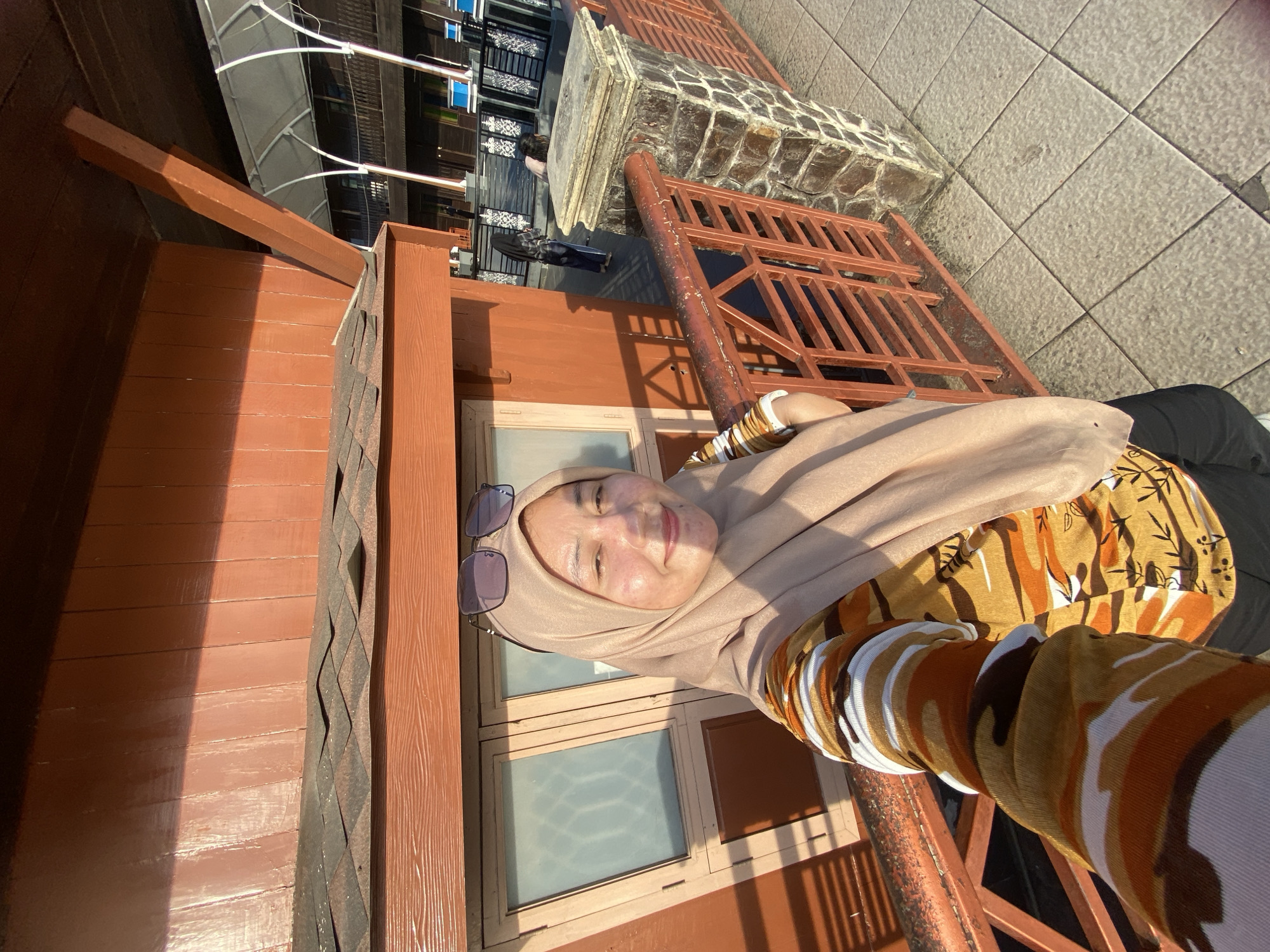 The image shows a woman wearing a tan hijab and a patterned shirt with orange, white, and brown camouflage-like designs. She is taking a selfie outdoors, with a wooden building with brown siding in the background. A railing made of metal bars and stone pillars is visible in the foreground. The woman is smiling and has sunglasses perched on her head. The environment appears to be a walkway or balcony with tiled flooring. In the background, other buildings and people are partially visible, suggesting a public area. The lighting indicates it is daytime, possibly late afternoon due to the long shadows. The location is described as being in the city of Banjarmasin, Indonesia.