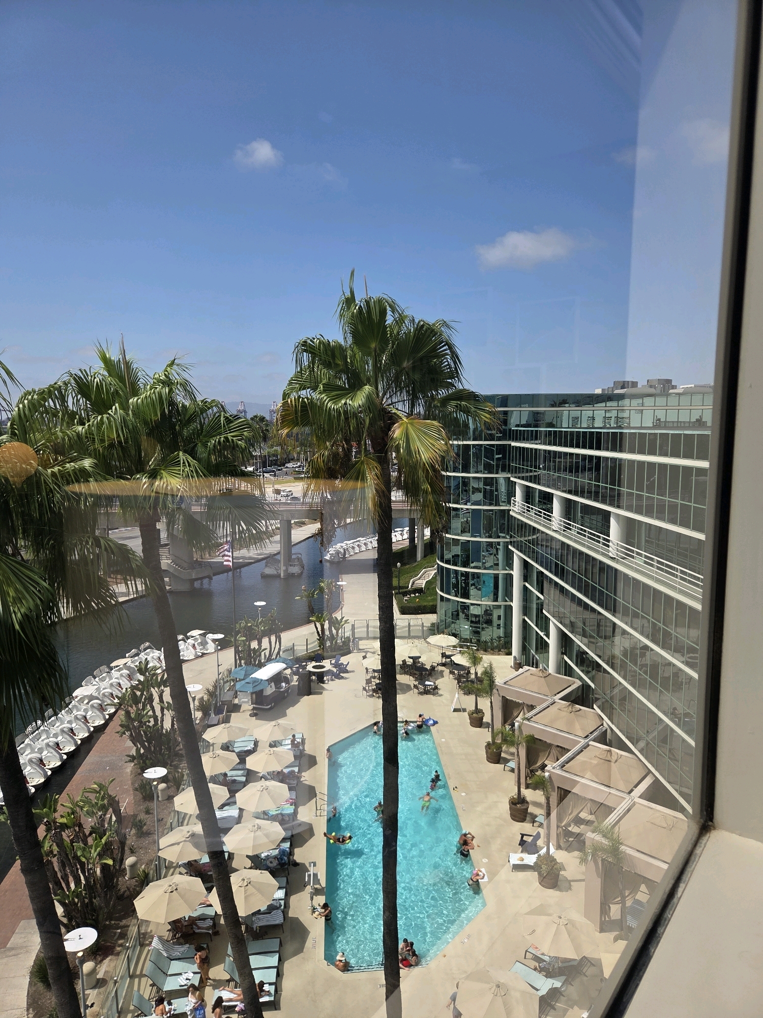 The image, taken from a high vantage point, offers a view of a hotel resort in Long Beach, California. A large, blue swimming pool is centrally located, with several people swimming and relaxing on lounge chairs scattered around it. Palm trees are abundant, with their fronds creating a tropical atmosphere.
To the left, a calm body of water, likely a marina or canal, is visible with several boats docked. A bridge spans across this waterway, with an American flag flying from a pole. On the right side of the image, a modern, glass-fronted building dominates the skyline, reflecting the blue sky and clouds. The building appears to be a hotel or office complex. The overall scene suggests a sunny day at a waterfront resort.