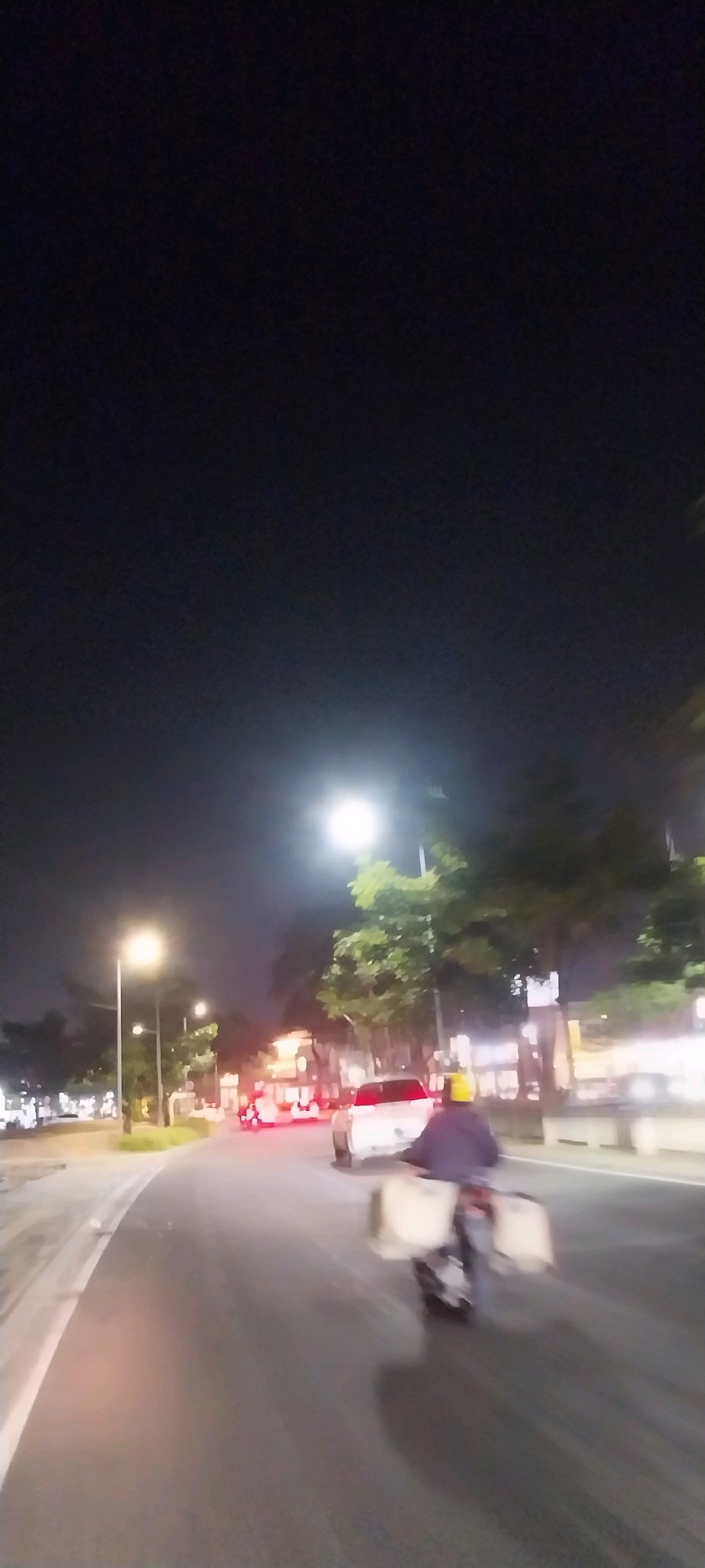 The image depicts a nighttime scene on a street in Medan, Indonesia. A person on a motorcycle, with a yellow helmet and a dark jacket, is traveling away from the camera. The motorcycle is equipped with two large, light-colored cargo boxes on the rear. In front of the motorcycle, a white car with its brake lights illuminated is also traveling in the same direction. Streetlights illuminate the road and the surrounding environment, which includes trees lining the street and buildings in the background. The overall scene is characterized by low light conditions and motion blur, suggesting the photo was taken from a moving vehicle.