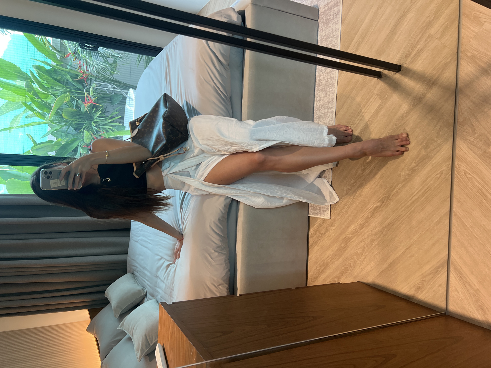A woman is taking a selfie in a bedroom. The woman has long dark hair and is wearing a black top and a white dress. She is holding a cell phone in her left hand and pointing it at the mirror in front of her. Her face is obscured by the phone. The woman has a Louis Vuitton bag next to her on the bed. The bedroom has a bed with gray bedding, a nightstand, and a window with a view of trees. There is a mirror on the wall next to the bed. The location is Unknown, Unknown.