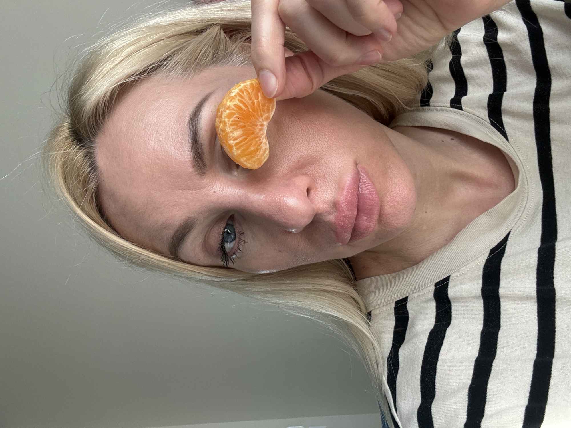 In this close-up, horizontal, and slightly angled shot, a woman with blonde hair is holding a segment of tangerine over her right eye. The tangerine segment is bright orange and curved, covering a portion of her eyelid and eyelashes. Her left eye, visible in the lower left of the frame, is open and has blue irises and visible eyelashes. Her skin appears to have a light makeup application, with a faint sheen. Her lips are slightly parted and have a pinkish hue.
The woman is wearing a striped shirt with thick black and cream-colored horizontal stripes. Only the upper portion of her torso and neck are visible. Her blonde hair is slightly out of focus around the edges of the frame.
The background is a plain, light gray wall. There are no distinct environmental cues to suggest a specific location or time of day beyond indoor lighting. The overall mood of the image is relaxed and possibly playful, due to the unusual placement of the fruit. No text is visible in the image.
