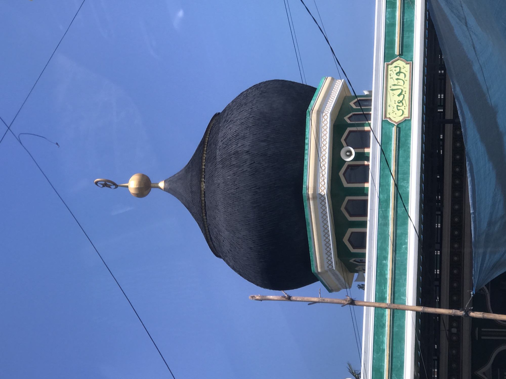 The image shows a mosque in Lhokseumawe, Indonesia. The mosque features a large black dome with a golden finial on top. A tall minaret adjacent to the dome is decorated with green and white stripes and Arabic script. Blue sky is visible in the background. Wires are strung overhead. Bamboo scaffolding is visible near the minaret.