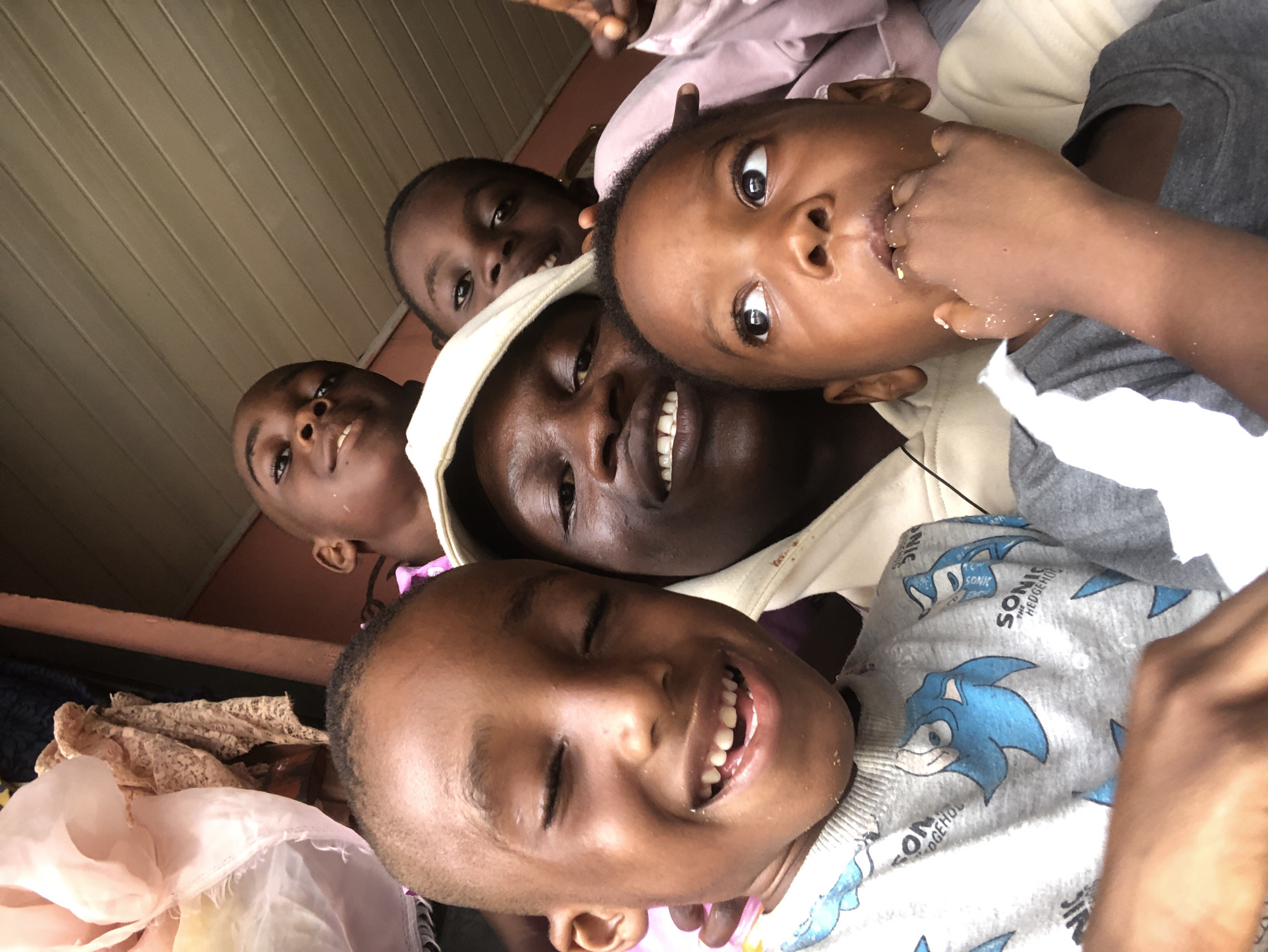 In the city of Ado, Nigeria, a group of five children and one adult are captured in a close-up, low-angle selfie. The subjects are positioned in the foreground, with the adult in the center, wearing a cream-colored hooded sweatshirt. To the adult's left, a child with closed eyes and a wide smile is wearing a gray sweatshirt featuring multiple prints of Sonic the Hedgehog characters. To the adult's right, another child is looking upwards with their hand near their mouth, appearing to be eating. Behind the adult, three other children are partially visible, smiling and looking towards the camera. The background consists of a wooden ceiling and a pink wall, with some draped fabric in the upper left corner. The overall impression is a spontaneous and joyous moment shared amongst a family or friends.