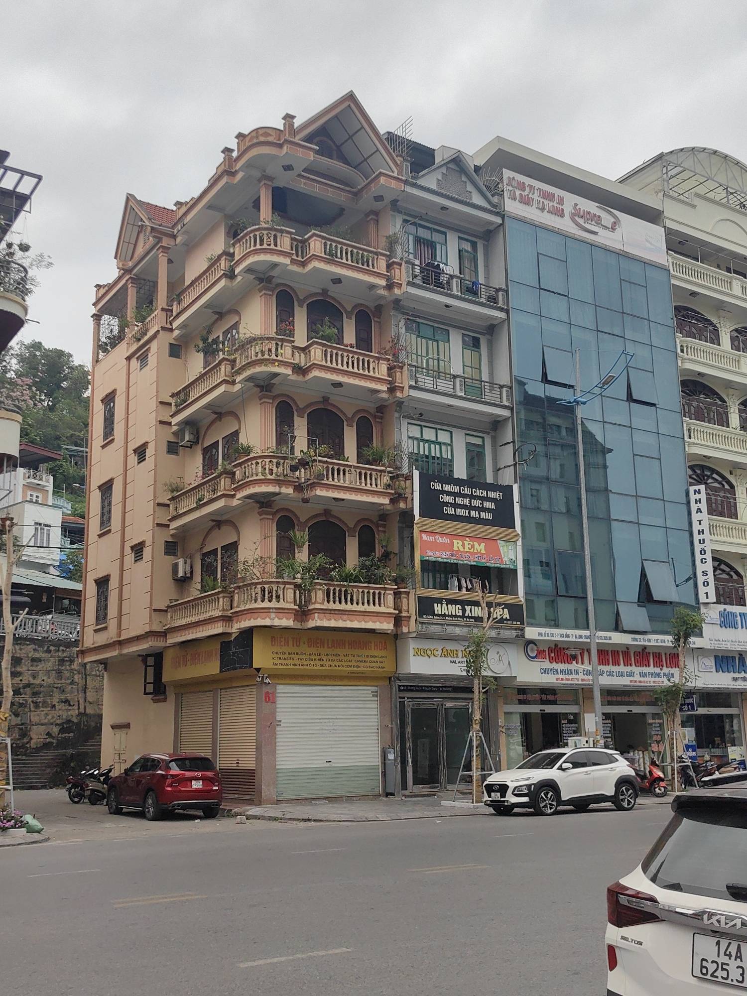 The image shows a street scene in Hong Gai Ward, Vietnam, featuring a row of buildings with diverse architectural styles. On the left is a multi-story building with arched balconies adorned with plants, a beige facade, and a traditional roof design. Adjacent to it is a narrower building with green-framed windows and a combination of signage for businesses, including "Nam Quan REM" and "HANG XIN PA." Next is a building with a modern glass facade reflecting the surroundings.
The street level features businesses with signage in Vietnamese, cars parked along the curb, and motorbikes nearby. Cars present are Kia Seltos, Mazda CX-5, and Hyundai Kona. The sky is overcast.