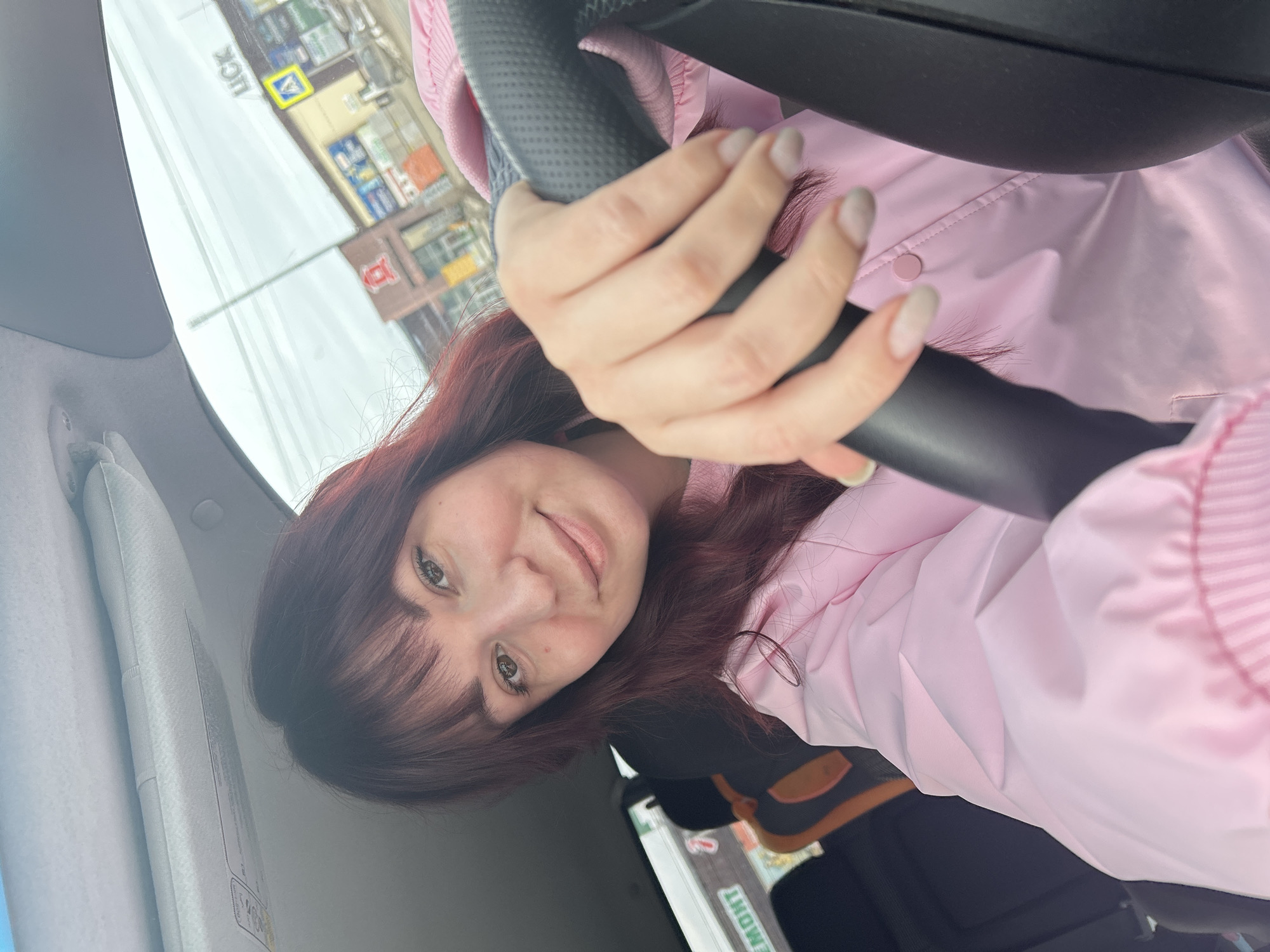 A woman with shoulder-length dark hair and bangs is in the driver's seat of a car. She has fair skin and is wearing a pink jacket. Her hands are on the steering wheel. The background shows a street scene in the city of Chaikovsky, Russia. A sign with the word "LUCK" is visible. There are buildings along the street. The sky is overcast.