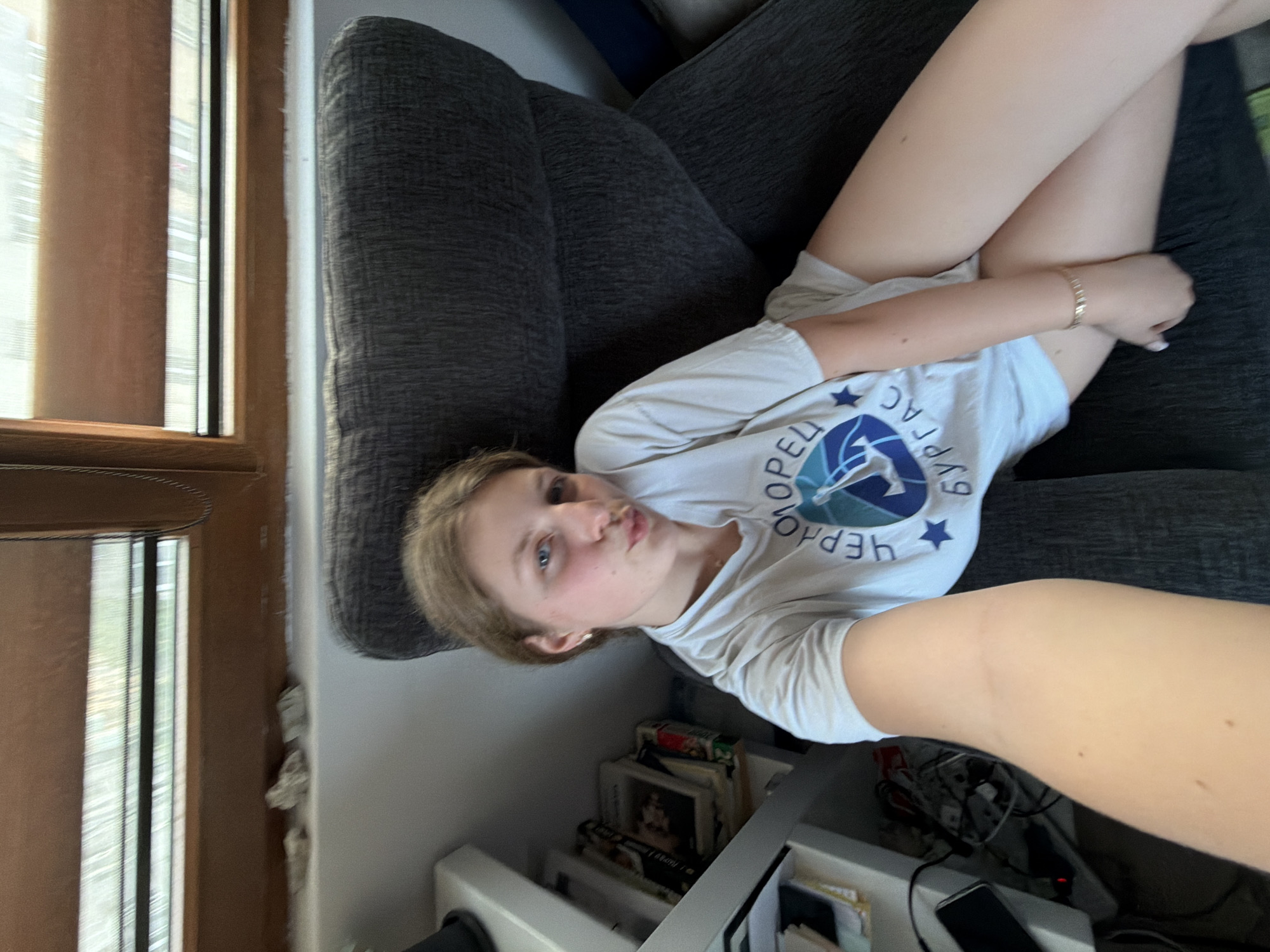 A young person is captured in a selfie, lying on their left side on a dark grey, textured sofa in an indoor setting. The individual has light brown hair, fair skin, and blue eyes, with a playful expression featuring slightly pursed lips. They are wearing a white t-shirt with a blue and white logo that includes a star and Cyrillic text. The visible text on the shirt reads "ЧЕРНОМОРЕЦ" (Chernomorets) and "БУРГАС" (Burgas), aligning with the provided location context of Burgas, Bulgaria. They are also wearing light-colored shorts and a delicate golden bracelet on their right wrist.
To the left of the sofa, a window with vertical blinds or shades allows natural daylight to enter the room, indicating it is daytime. Below the sofa, a white shelving unit is visible, filled with books, small objects, and some tangled cables, suggesting a lived-in space. A corner of a phone or similar electronic device can be seen in the lower right foreground, reinforcing the selfie nature of the photograph, likely held by the subject. The overall mood is casual and relaxed.