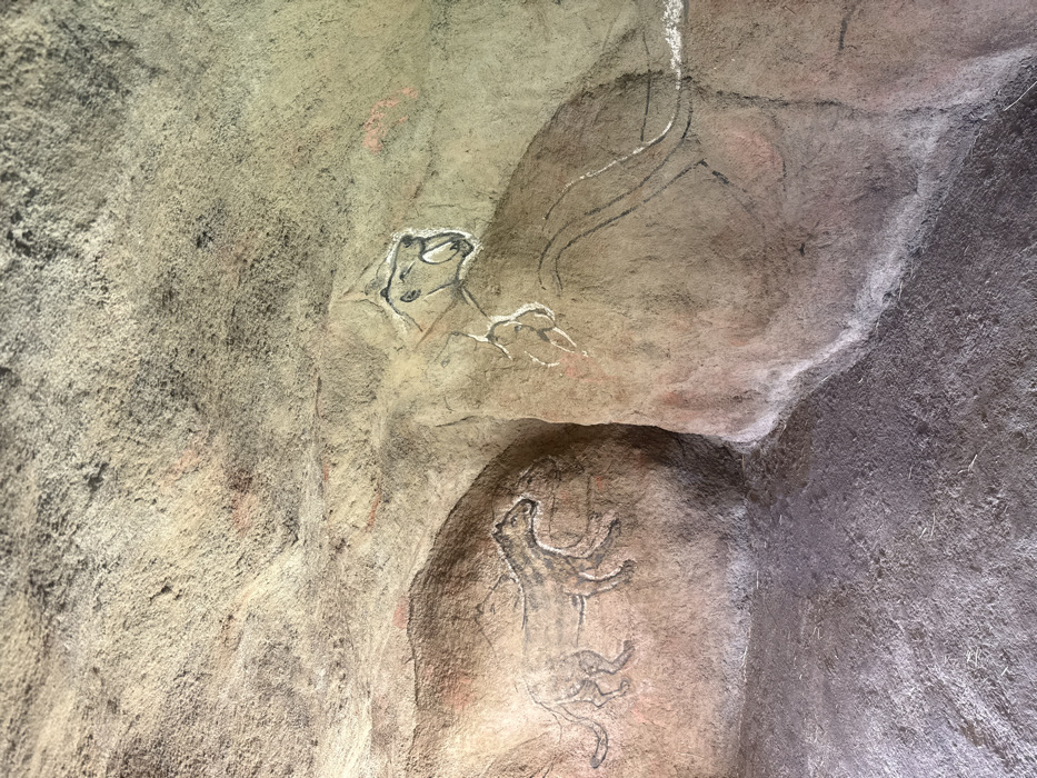 The image depicts a simulated cave interior, consistent with an educational exhibit, in the City of Zagreb, Croatia. The rough, undulating walls, varying in color from light beige and grey to reddish-brown, are adorned with two large-scale, stylized representations of prehistoric animals, rendered in a manner emulating Paleolithic cave art. The floor is dark grey and granular, simulating a cave ground. On the left wall, a quadruped animal, possibly a large feline or canid, is outlined in black with faint internal stripes, appearing to stride to the right. On the right wall, a larger predatory animal with an open mouth, prominent saber-like fangs, and visible claws is depicted in black outline with white highlights, conveying an aggressive stance. No human subjects or actions are present beyond the artwork itself. This scene is likely an installation within an educational institution, such as the Croatian Natural History Museum (Hrvatski prirodoslovni muzej), in the City of Zagreb.