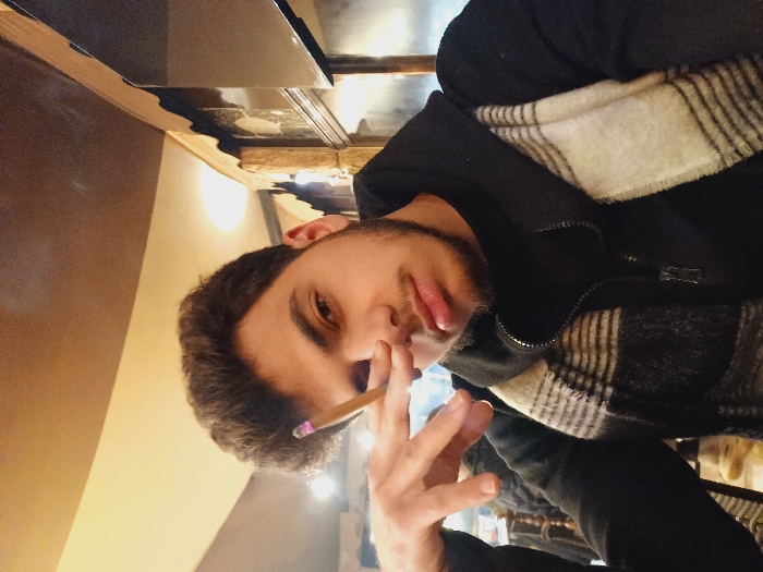 A young man is pictured in what appears to be an indoor setting, possibly a café, with warm lighting. He's holding a lit cigarette or joint in his right hand, gesturing slightly with his fingers. The man has dark hair, a light beard, and is wearing a dark jacket with a plaid scarf visible at the collar. He looks directly at the camera with a somewhat intense gaze. There is some kind of counter with a reflective top visible behind the subject. The image is rotated counterclockwise.