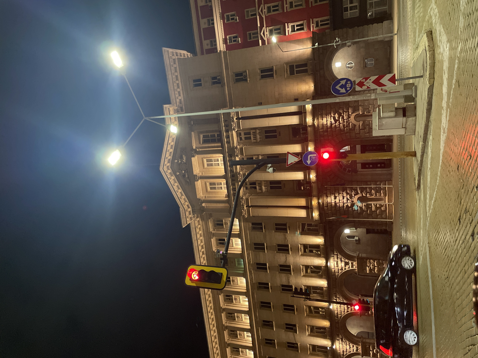 The image depicts a nighttime scene in Sofia, Bulgaria. The backdrop is dominated by an imposing, illuminated building with a classical architectural style. The building features multiple floors, numerous windows, and ornate detailing. A streetlight with two arms shines brightly on the left side of the image. Traffic lights are visible at different points, all displaying red signals. A black car is parked on the right side of the street. Road signs are visible near a traffic light on the right side of the image. The street appears to be paved with light-colored stones or bricks. The sky is dark, adding to the nighttime atmosphere.
