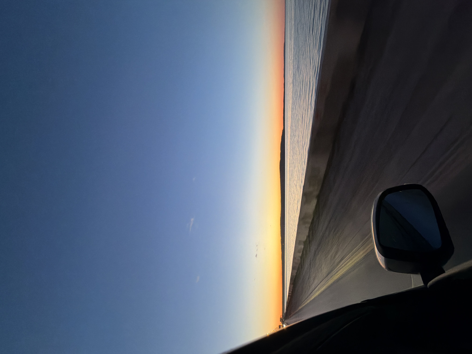 The image shows a view from inside a car, possibly driving alongside a body of water at sunset. The sky transitions from blue to orange as it approaches the horizon. A car's side mirror is visible. The location cannot be confidently determined to be Chaikovsky, Russia.