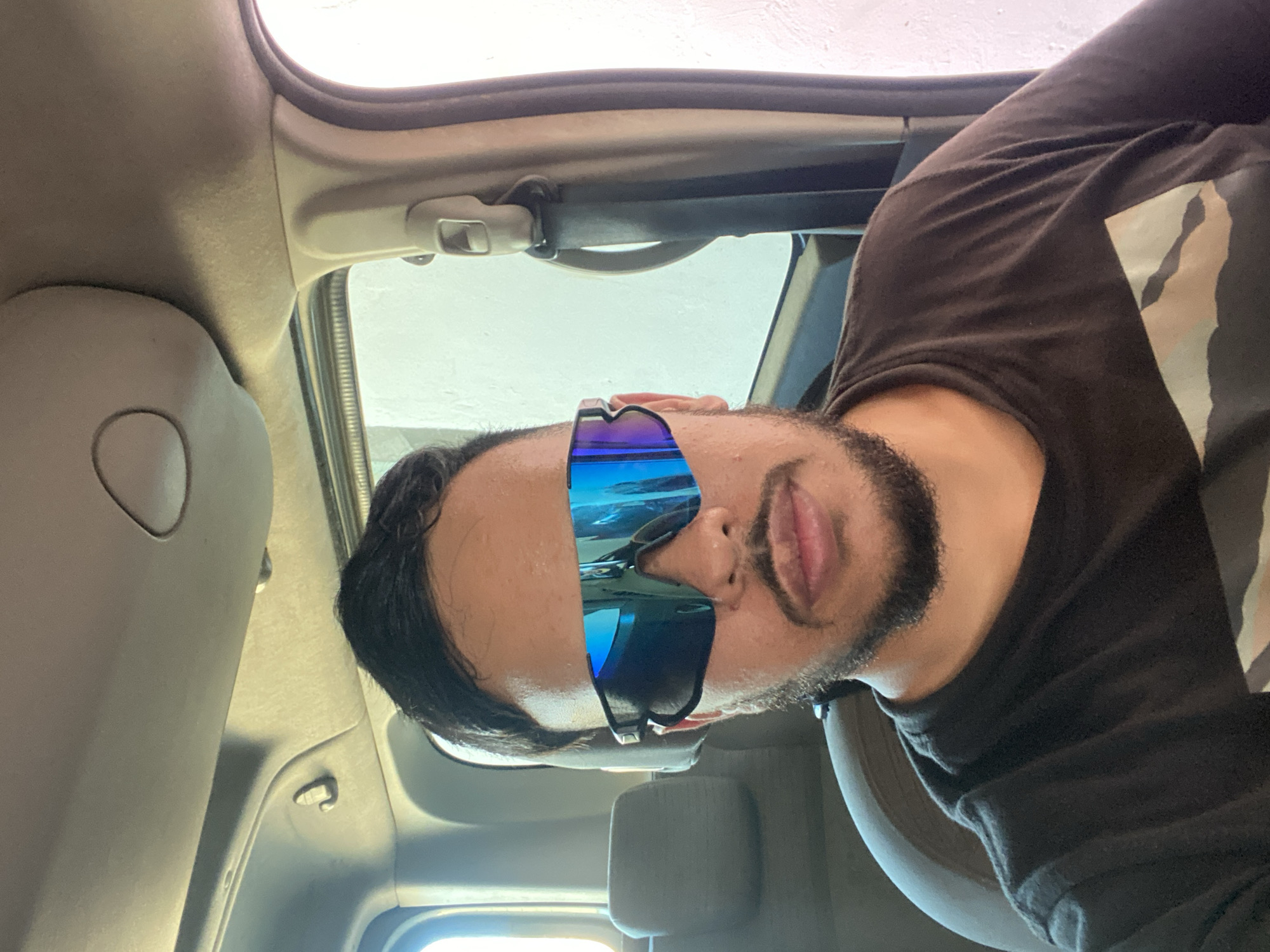 A man with dark hair, a beard, and a mustache is sitting in the passenger seat of a car. He is wearing a black t-shirt and large, reflective blue sunglasses. The interior of the car is visible, including the ceiling, a seatbelt, and part of the headrest of the seat in front of him. The background is out of focus, but appears to be a light-colored wall and a window. The lighting suggests it is daytime.