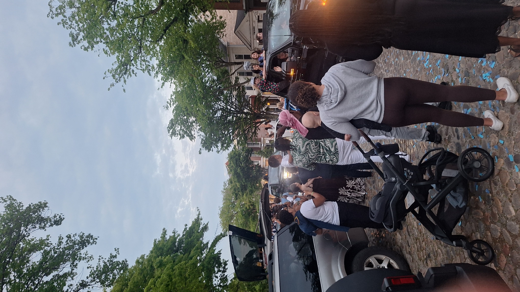 In a cobblestone street lined with trees and houses, a group of people are gathered around vehicles, with some standing on top of a black SUV. The sky is overcast, and the scene appears to be outdoors in a residential area. Several individuals are waving, and a baby stroller is present on the ground. There are scattered blue pieces of paper on the cobblestones. The overall impression is that of a celebratory or festive event.