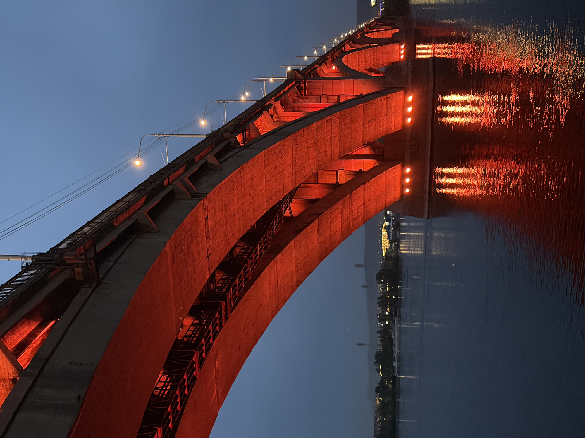 The image shows a view of the Kommunalny Bridge over the Yenisei River in Krasnoyarsk, Russia, illuminated by red lights at dusk. The bridge's concrete arches are prominently lit from below, casting a vibrant glow onto the water's surface, where the reflections create streaks of light. The dark blue sky is visible above the bridge, with a few streetlights lining its upper structure. In the distance, the city skyline is discernible with scattered lights and trees. A white boat is seen on the river. The overall scene captures the bridge at night, highlighting its architecture and the ambient lighting.