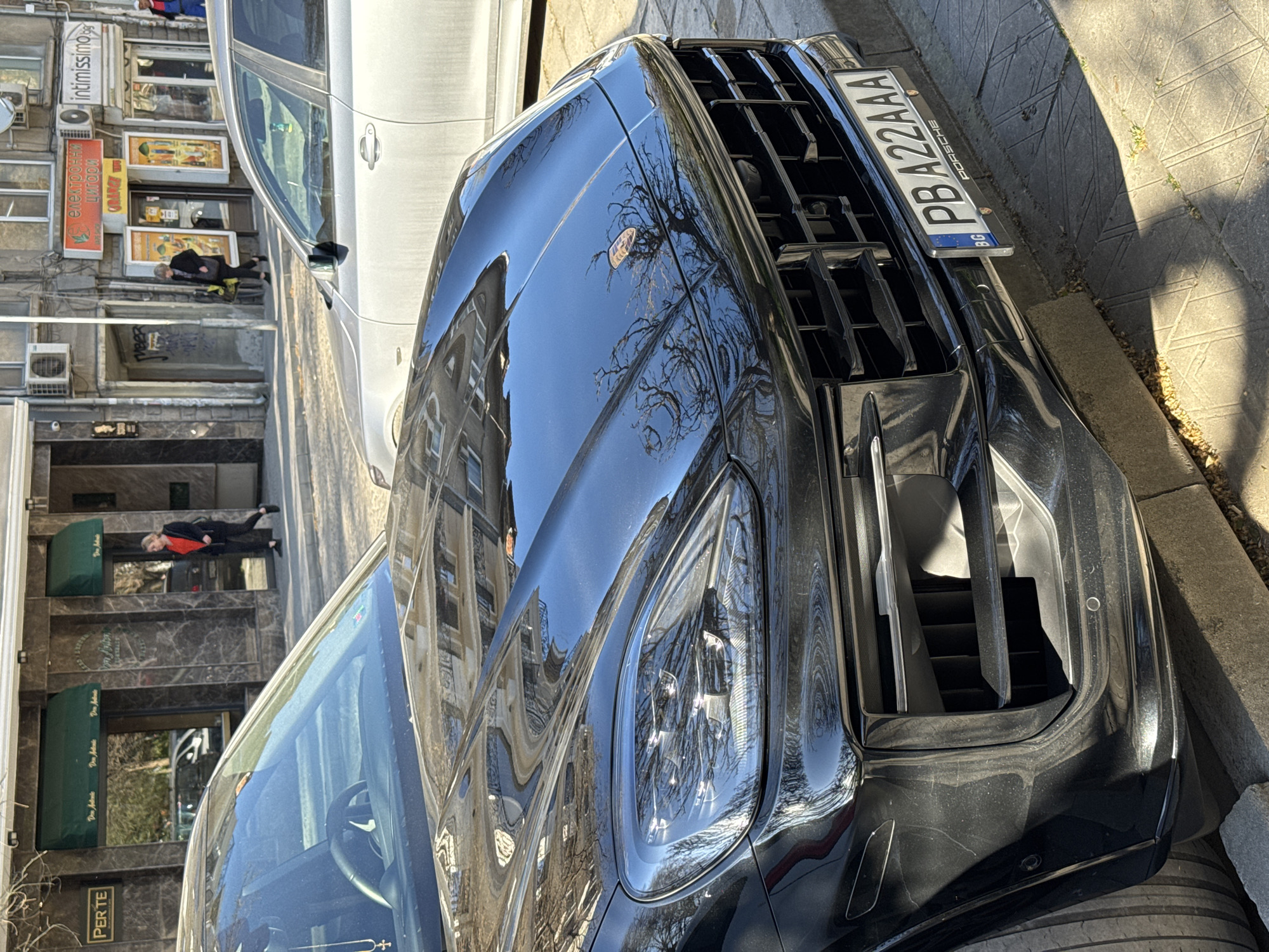 The image shows a black Porsche car parked on a street. The reflection of a building is visible on the car's hood. A license plate reading "PBA242AA" is affixed to the vehicle. In the background, buildings and pedestrians are visible. A sign for "Intimissimi" and "Електроуреди" can be seen. The reflection on the car also show the "Perte" store. It appears to be a street in Plovdiv, Bulgaria.