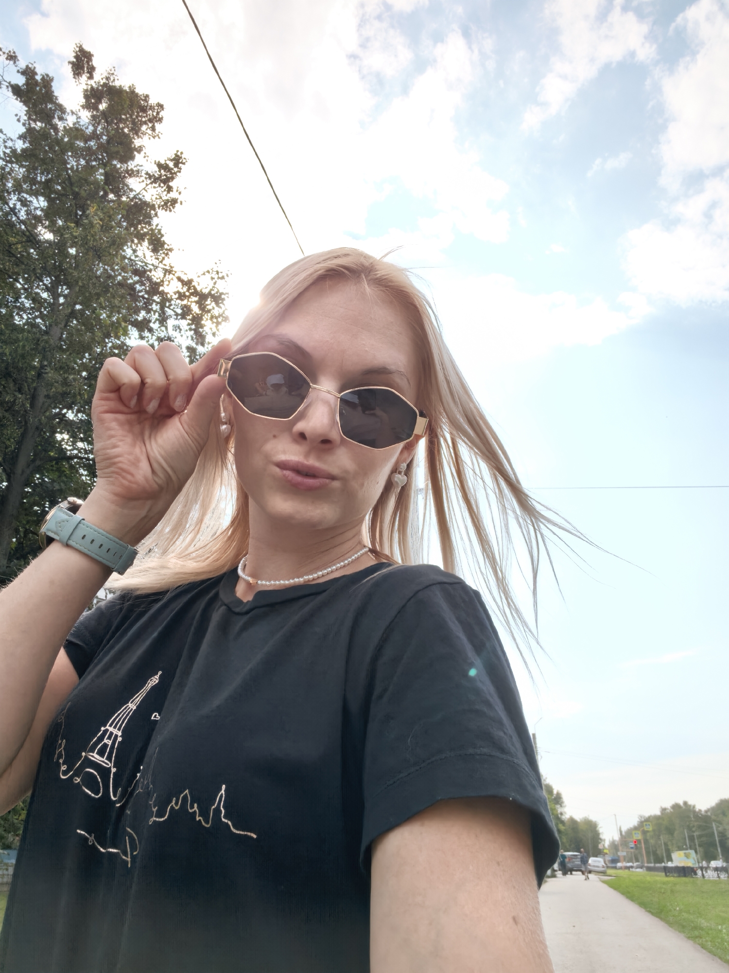 A blonde woman, wearing sunglasses, a black t-shirt with an Eiffel Tower graphic, a pearl necklace, and a light blue smartwatch, poses for a selfie outdoors. She is holding her sunglasses with her left hand. Her hair is blowing in the wind. The background shows a cloudy sky, trees, a sidewalk, a street with cars, and buildings in the distance. The specific location in Sterlitamak, Russia is not identifiable from the image.