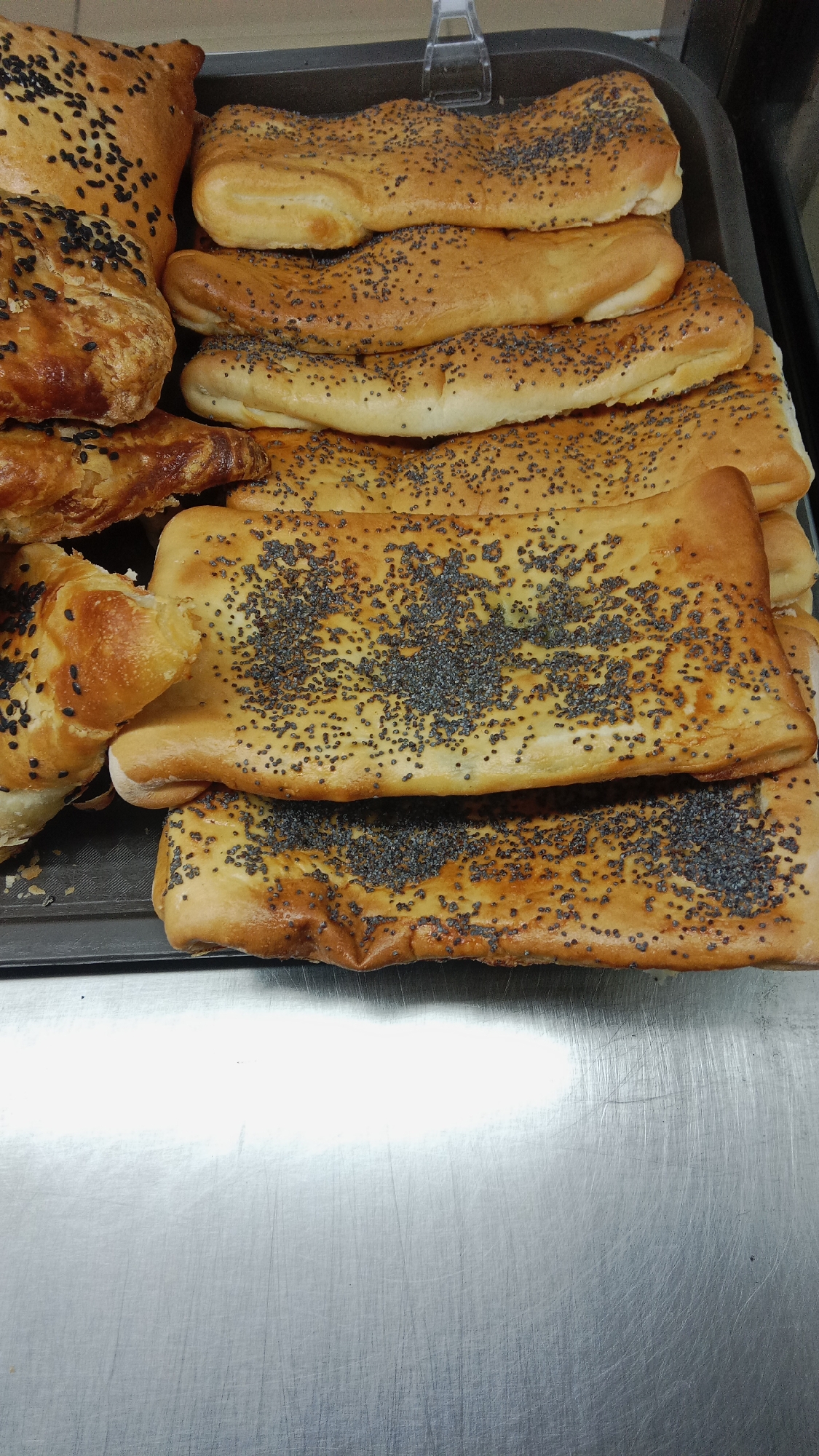 A tray of freshly baked bread is displayed on a metal counter. The bread appears to be of various shapes and sizes, with some pieces topped with poppy seeds and others with black sesame seeds. The bread is golden brown and appears to be soft and fluffy. The metal counter is clean and shows some signs of wear. The overall setting suggests a bakery or a food service establishment in Petropavl, Kazakhstan.