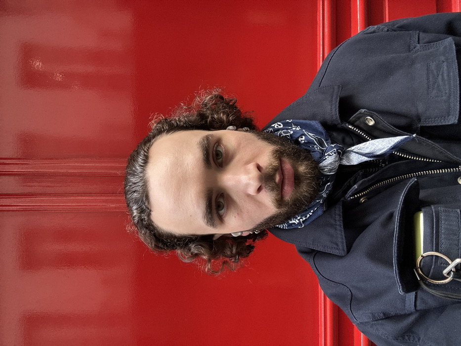 The image shows a headshot and upper torso of a man positioned against a vibrant, glossy red background, which appears to be a door or wall panel with horizontal moldings. The man is looking directly at the camera with a neutral expression.
He has dark, curly hair that appears to be well-groomed, and a neatly trimmed dark beard and mustache. His eyes are a striking light color, appearing greenish or hazel. He wears small, silver-colored stud earrings in both ears and has small white earbuds in each ear.
The man is dressed in a dark blue or navy jacket with a structured collar. The jacket has visible zippers, including a main one down the front and possibly others on pockets, and silver-colored snap buttons on the lapel area. Around his neck, he wears a blue bandana with a white, intricate pattern, likely paisley, tied in a knot.
In a pocket of his jacket, on the right side of the image (viewer's left), a light green or yellow object, possibly a phone or a card holder, is visible, along with a keyring holding a couple of keys or rings. The background's glossy finish reflects some light, creating subtle highlights on its red surface. The lighting appears soft and even, highlighting the man's features.