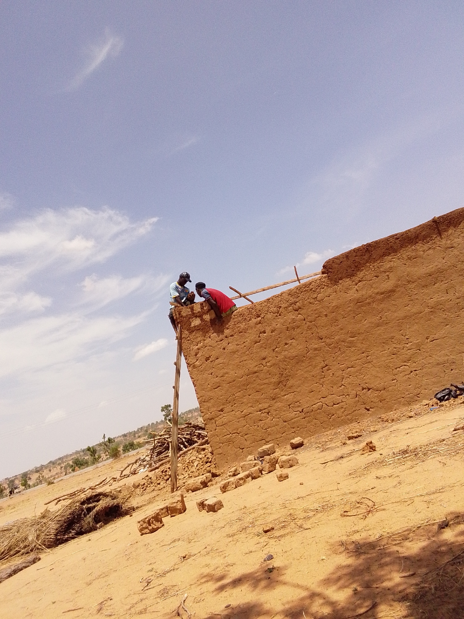Two men are on the roof of an unfinished mud building. One man in a light blue shirt and black cap is working, while the other man in a red shirt looks on. A crude wooden ladder leans against the building. The surrounding environment is arid, with a sandy ground and sparse vegetation. Scattered debris and building materials are visible. The photograph was taken in Mulke Barikin Sayejo, Nigeria.