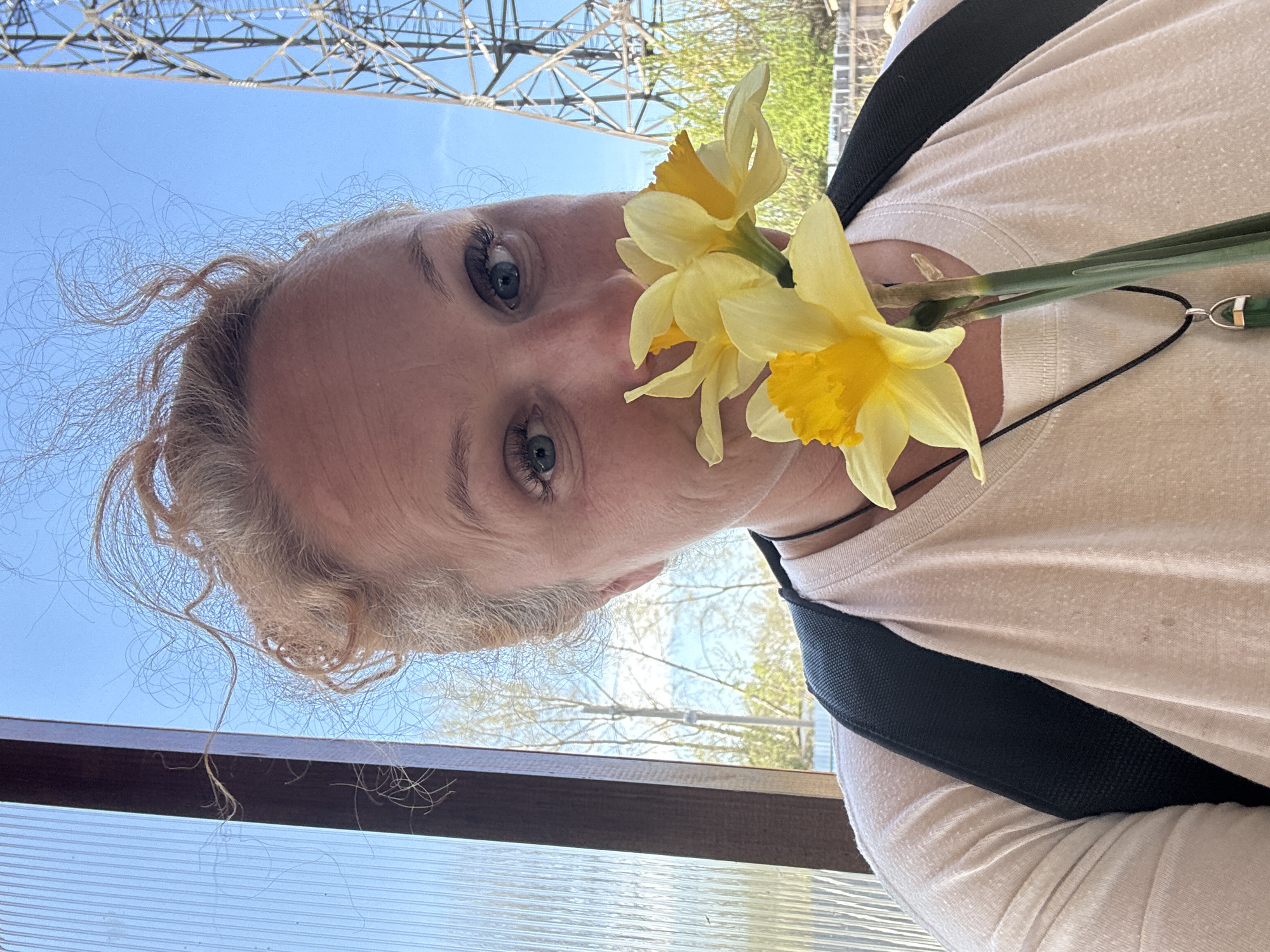 A woman with fair skin, light hair, and blue eyes holds a bouquet of three yellow daffodils close to her face. The photograph is taken from a low angle, looking up at the woman. She wears a light-colored shirt and a black strap crosses her chest, possibly from a backpack or bag. A necklace with a green pendant is visible. Above her, there are overhead power lines against a blue sky, with trees and structures visible in the background. The location appears to be Chaikovsky, Russia based on geolocation evidence, though I am unable to confirm this with a confidence level above 70%. The photograph seems to be taken from within a structure with a window or glass enclosure.
