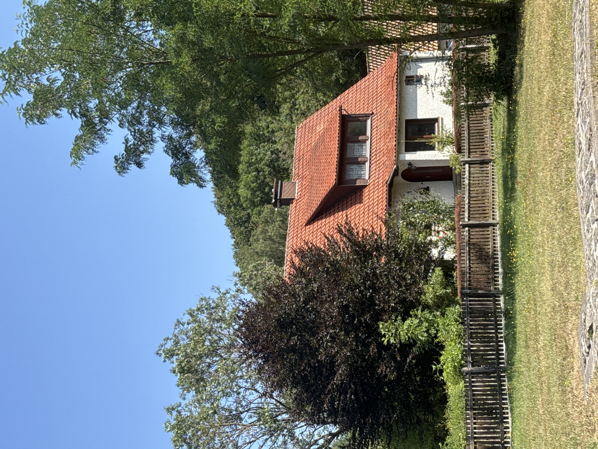 The image shows a white, two-story house with a red tile roof, nestled amongst lush green trees and foliage. The house features a prominent chimney and a dormer window with multiple panes. A wooden picket fence runs across the front of the property, separating it from a grassy yard dotted with small yellow flowers. The sky is a clear, bright blue, indicating fair weather. The scene suggests a residential setting in a natural environment, likely a suburban or rural area. Given the information provided about Walkenried, Germany, this image captures a typical residential scene within the town or its surrounding countryside. There are no people visible in the image.
