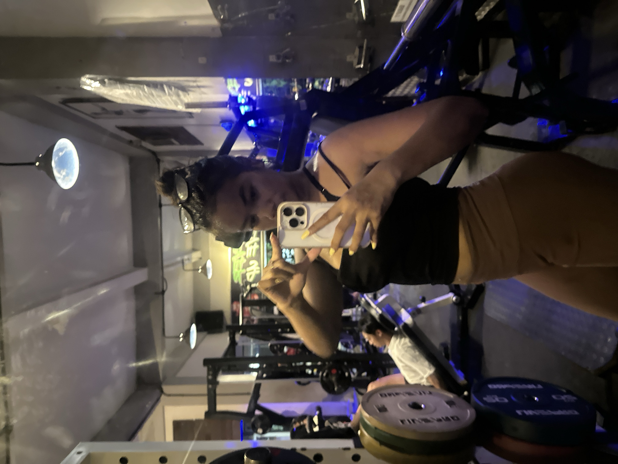 A young woman with dark hair pulled into a bun and sunglasses perched on her head is taking a selfie in a gym in Denpasar, Indonesia. She is wearing a black tank top and tan leggings, with bright yellow nail polish. She is holding a smartphone with a white case. In the background, a person is lying on a weight bench, and various gym equipment, including weight plates and racks, are visible. The gym appears to be dimly lit with overhead lights and blue accent lighting.
