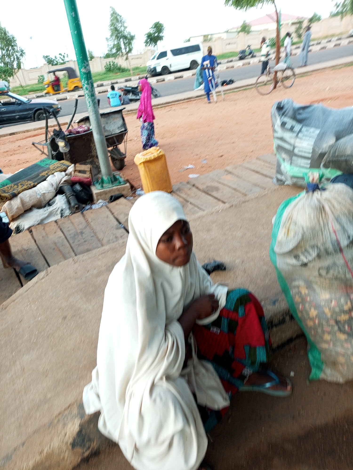 The image depicts a street scene in Gombe, Nigeria, with a young girl wearing a white hijab and a colorful skirt sitting on concrete steps. She is looking to her right with a focused expression. In the background, a yellow auto-rickshaw is visible, as well as other people, vehicles, and various objects like sacks and a yellow barrel. The environment appears to be a public area with a road and a sidewalk or pathway. The lighting suggests it is daytime. The overall impression is of a casual, everyday scene in a bustling urban environment.