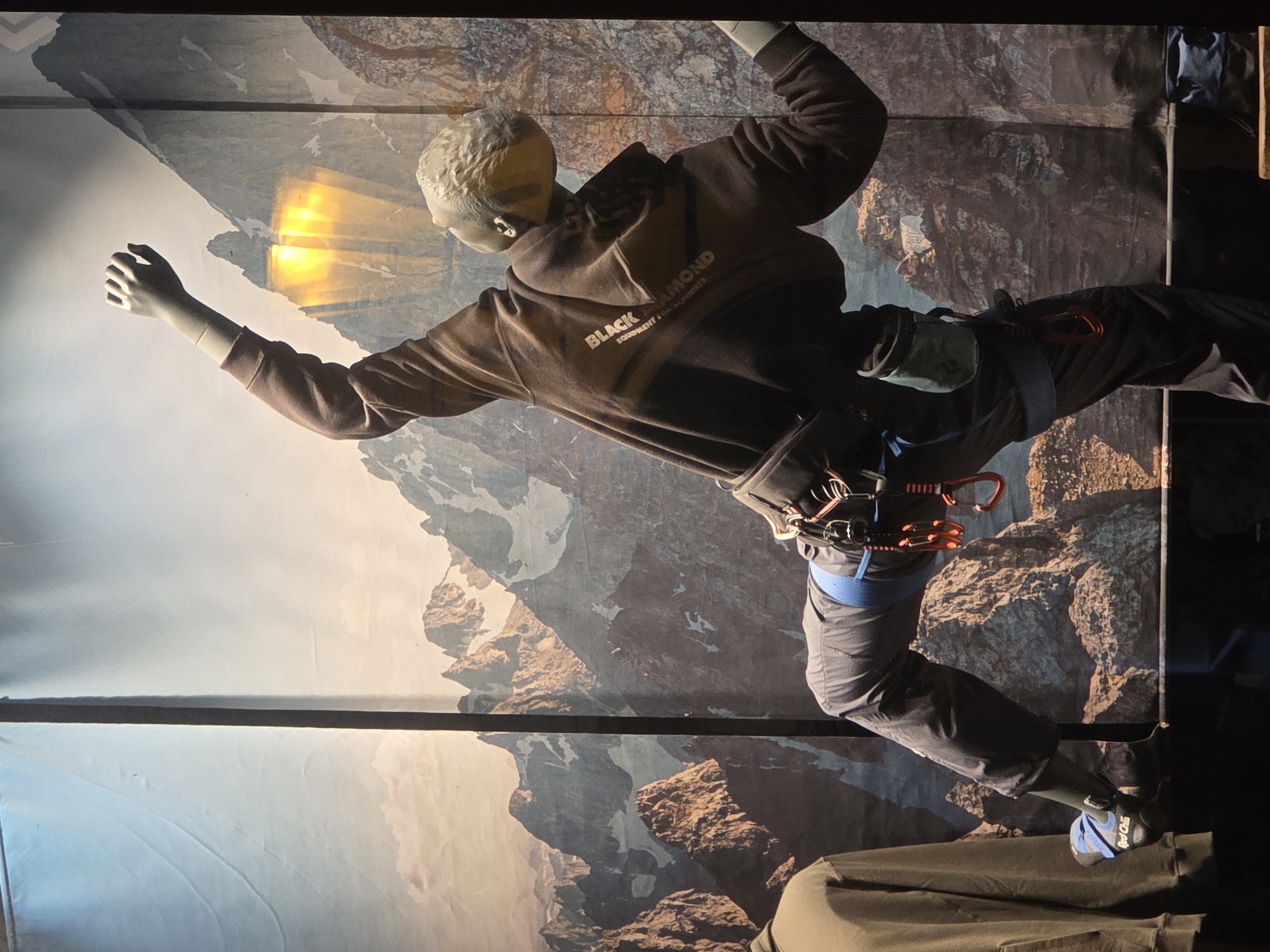 The image shows a mannequin posed as a rock climber against a backdrop of a mountain range. The mannequin is wearing a Black Diamond hoodie, climbing pants, a climbing harness, and climbing shoes. The mannequin is in a climbing pose with one arm extended and the other reaching up. The background is a photographic backdrop of a mountain range. The location is unknown.