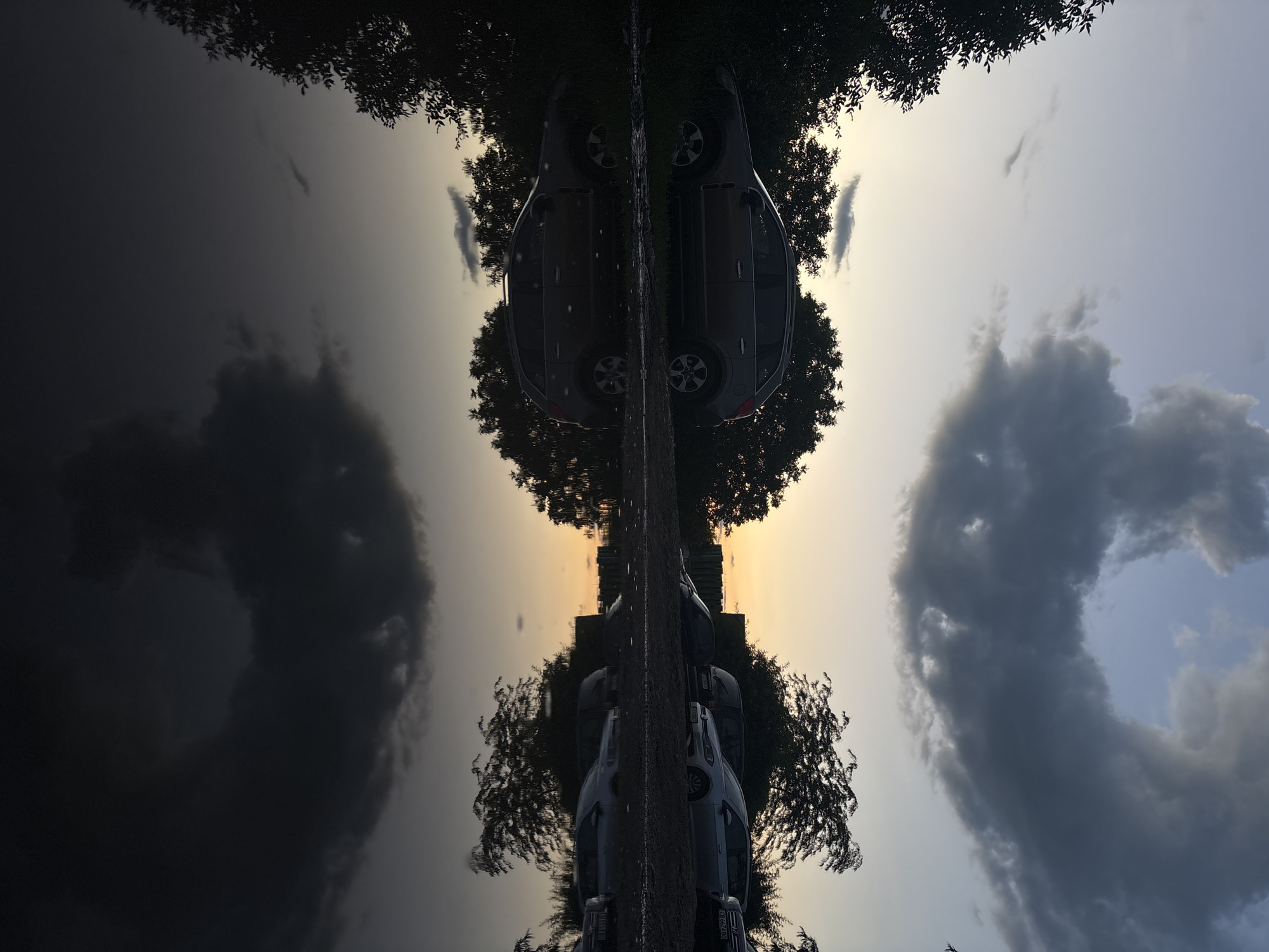 The image depicts a car parked on a dirt road next to a large puddle, reflecting the sky and trees. The sky is a twilight hue with dramatic, dark clouds and a sliver of orange on the horizon. Several other vehicles are visible in the background, parked on a gravelly area. The scene appears to be an outdoor parking area, possibly on the outskirts of a city, given the natural elements and the mix of vehicles. The lighting suggests it is either dawn or dusk. There is no specific indication of the location being in Sofia, Bulgaria, within the image itself.
