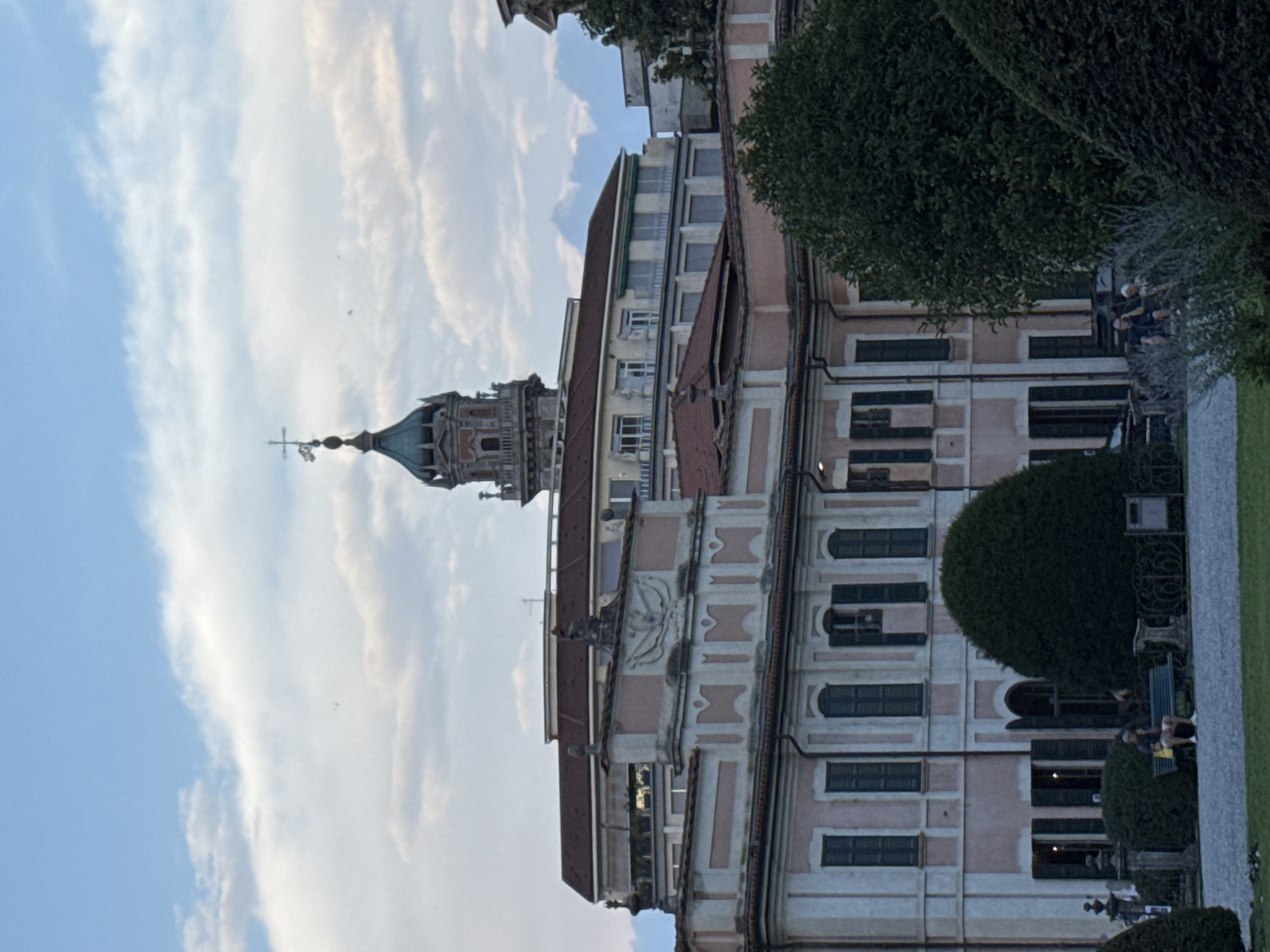 The image captures a view of the Palazzo Estense in Varese, Italy, with a cloudy sky overhead. In the foreground, a man is seated on a bench in front of a large, dark green, rounded bush. Behind him, the pale pink facade of the Palazzo Estense is visible, featuring numerous windows with dark shutters and architectural details. A prominent dome with a cross on top rises above the building in the distance. The ground is covered with gravel and grass. The lighting suggests it is either late afternoon or early evening.