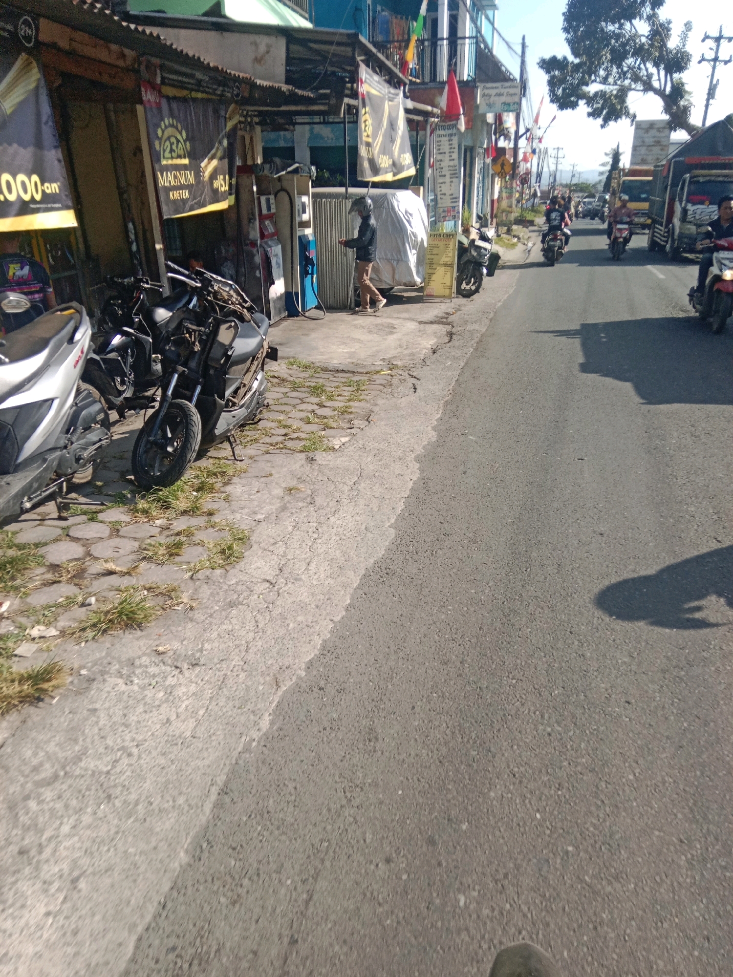 The image shows a street scene in Kertek, Indonesia, likely a commercial area. In the foreground, a silver scooter is parked on the left, and a black scooter is parked next to it. Both scooters are on a paved area with hexagonal tiles and scattered grass. Behind the scooters are several shops with awnings. One shop has a banner advertising "MAGNUM KRETEK" with a price of "Rp15.000". A person wearing a black jacket and helmet is walking away from a blue and red structure, possibly a fuel dispenser, towards the shops. Further down the street, several motorcycles are in motion, and a truck is visible on the right side of the road. The sky is clear and blue, and there is a large tree on the right side of the image.