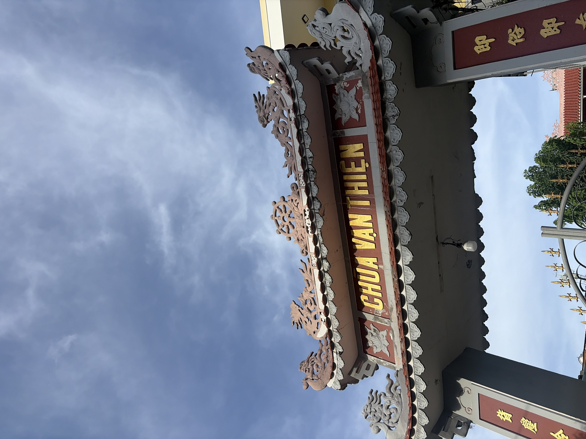 The image shows a close-up, low-angle shot of the ornate gateway of Chua Van Thien pagoda in Da Nang, Vietnam. The gateway is decorated with intricate carvings of dragons and lotus motifs, painted in shades of pink, grey, and gold. Golden Vietnamese characters are inscribed on the gateway, reading "Chua Van Thien." The sky above is a pale blue with wispy clouds. On the left and right pillars of the gateway, there are more golden Vietnamese inscriptions. A bare light bulb hangs from the ceiling of the gateway. Trees and part of the pagoda roof are visible in the background.