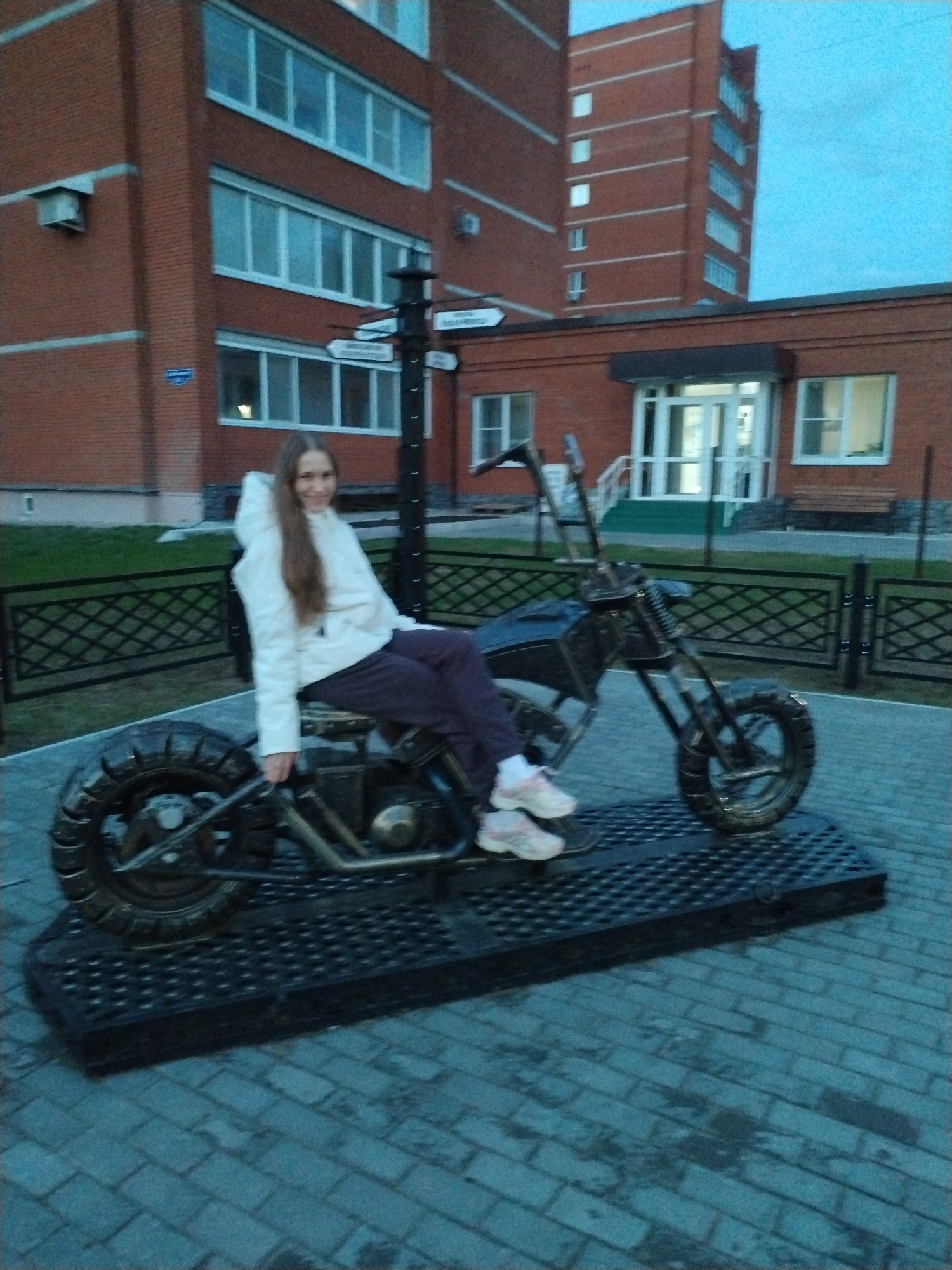 A medium shot shows a young woman sitting on a metal motorcycle sculpture outdoors in the city of Chaikovsky, Russia. The woman is wearing a white hooded jacket and dark pants, and pink sneakers. She smiles and faces the camera. The motorcycle sculpture is black metal with broad tires and sits on a black metal platform. Behind the platform is a low, black metal fence. Beyond that, a patch of green grass and two buildings made of red bricks. A multi-directional signpost is behind the motorcycle, next to the lower brick building. The ground is paved with blue-grey bricks. The sky is overcast and the lighting is subdued, suggesting the image was taken during twilight or under heavy cloud cover.