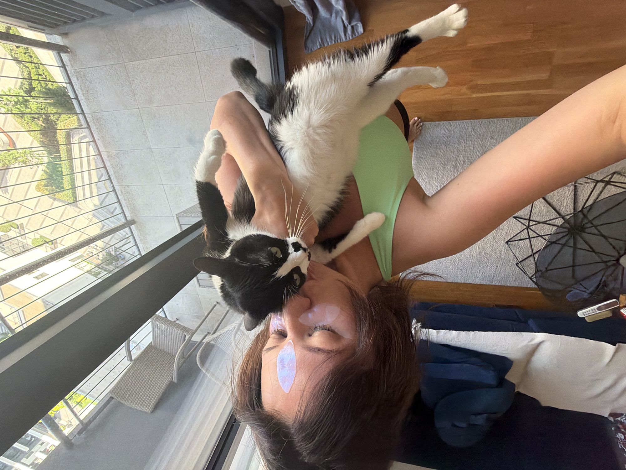 In this selfie, a woman is embraced by her black and white cat, who is nuzzling her cheek. The woman is wearing a light green tank top and has iridescent eye patches on her forehead. The scene appears to be indoors, near a large window or balcony. Outside, a cityscape with greenery and buildings is visible, suggesting an urban environment. The lighting implies it is daytime, possibly late morning or early afternoon. The woman's eyes are closed, and her expression, along with the cat's affectionate gesture, conveys a sense of warmth and affection. In the background, a wooden floor and a rug are visible, along with some furniture like a chair and what seems to be a folded blanket or bedding.