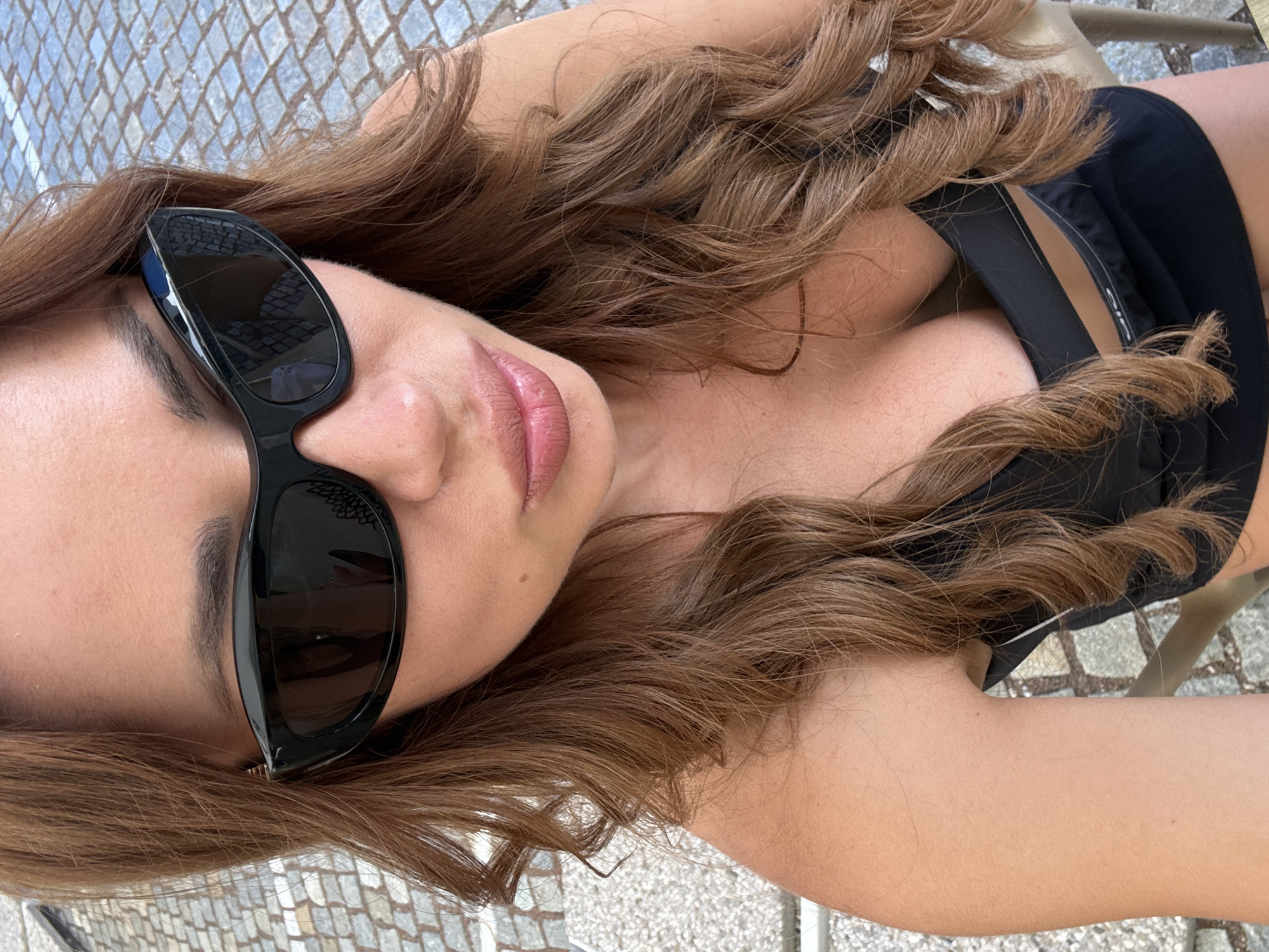 A medium shot captures a woman with long, wavy, reddish-brown hair and fair skin. She is wearing large, black, wraparound sunglasses and a black top that reveals cleavage. Her lips are parted slightly and appear to have a pinkish-toned lipstick. The background shows a cobblestone surface, suggesting an outdoor, possibly urban or village setting, with some out-of-focus elements suggesting furniture or a chair. The image is a selfie, taken from a high angle looking down. The setting is Colfosco, Italy.