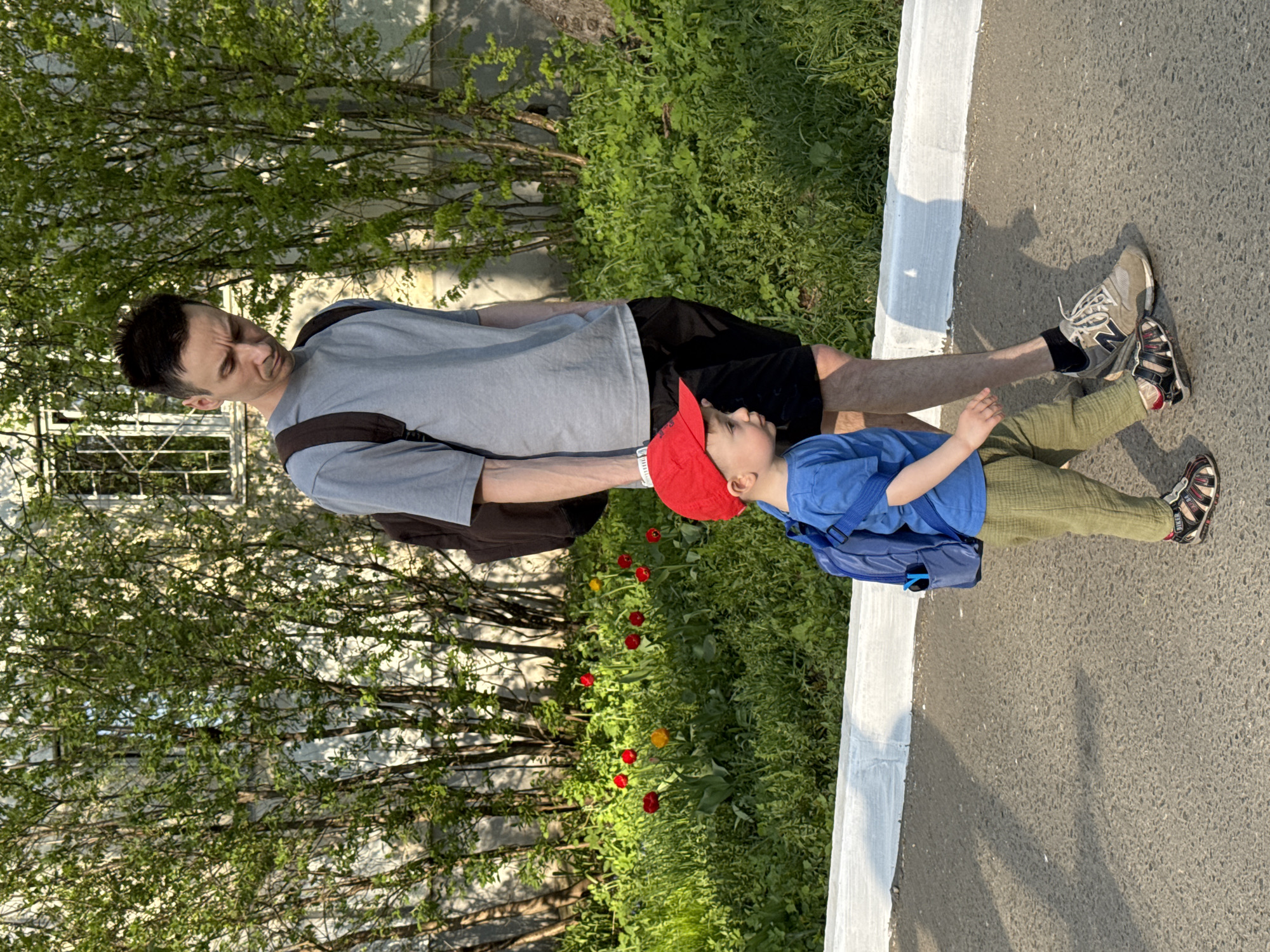 A man and a young child are standing on a paved surface next to a white curb, possibly a sidewalk, near foliage. The man is wearing a grey t-shirt, black shorts, and grey running shoes with black socks. He has short dark hair. He is wearing a backpack and is looking down towards the child. The child is wearing a red baseball cap, a blue t-shirt, green pants, and red and black sandals. The child also has a backpack. There are trees and grass growing in the background. Some red and yellow tulips are also visible. The location appears to be an urban or suburban area, potentially in Izhevsk, Russia.